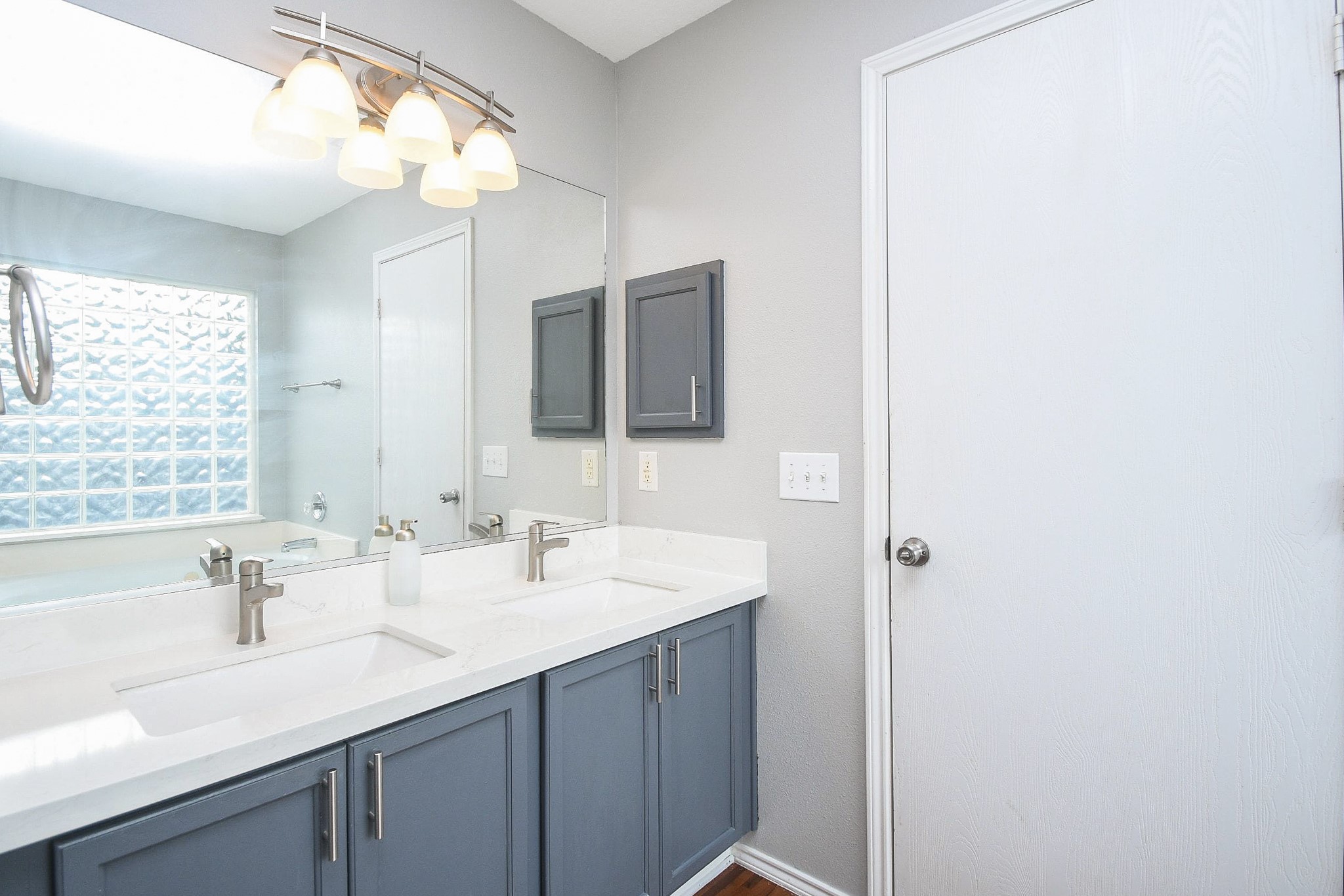 Elegant bathroom featuring a double vanity with sleek faucets, modern lighting, and ample cabinet storage, complemented by a stylish glass block window for natural light.