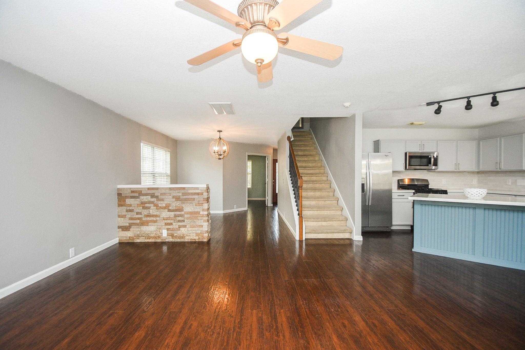Spacious open-concept living area with rich wood floors, modern kitchen featuring stainless steel appliances, and a stylish breakfast bar, all complemented by a cozy brick accent wall.