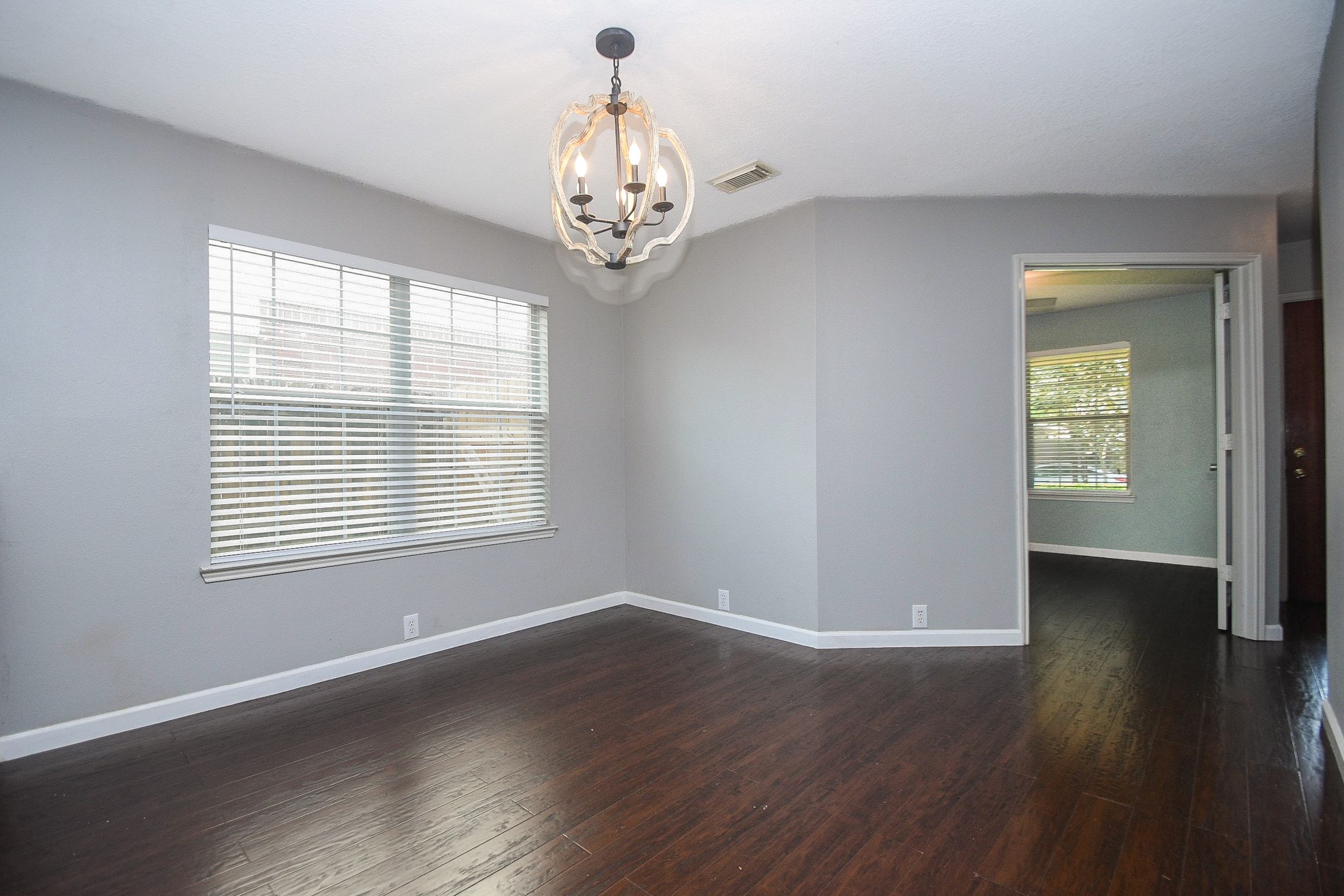 Bright and inviting room featuring rich hardwood floors, a modern chandelier, and ample natural light from large windows, with a doorway leading to an adjacent space.