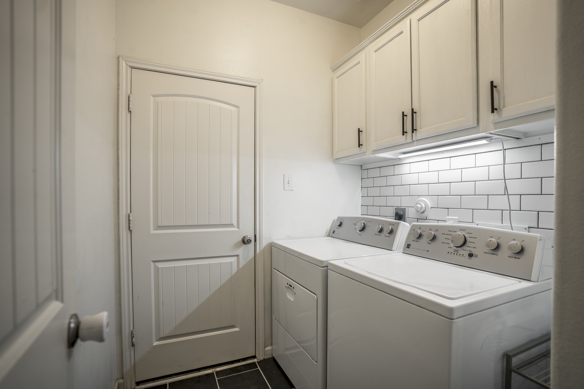 Back downstairs, the utility room keeps laundry and storage tucked into their own space just off the main hall. Built-in cabinetry, under-cabinet lighting, and a subway tile backsplash give the room a finished look, while new tile flooring keeps it practical for everyday use. The door ahead leads directly into the garage, making this an easy drop zone for coming and going.