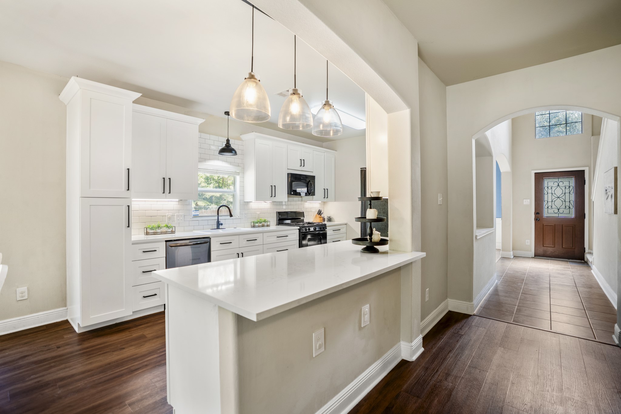 This angle gives you one of the best looks at the remodeled kitchen, with white cabinetry, black hardware, modern pendant lighting, and the breakfast bar opening the space up for casual seating and serving. Straight ahead, the hallway leads back toward the foyer and front of the home, while the breakfast area sits just behind and to the left of where youre standing here. Its a clean, connected layout that keeps the kitchen central to both everyday meals and entertaining.