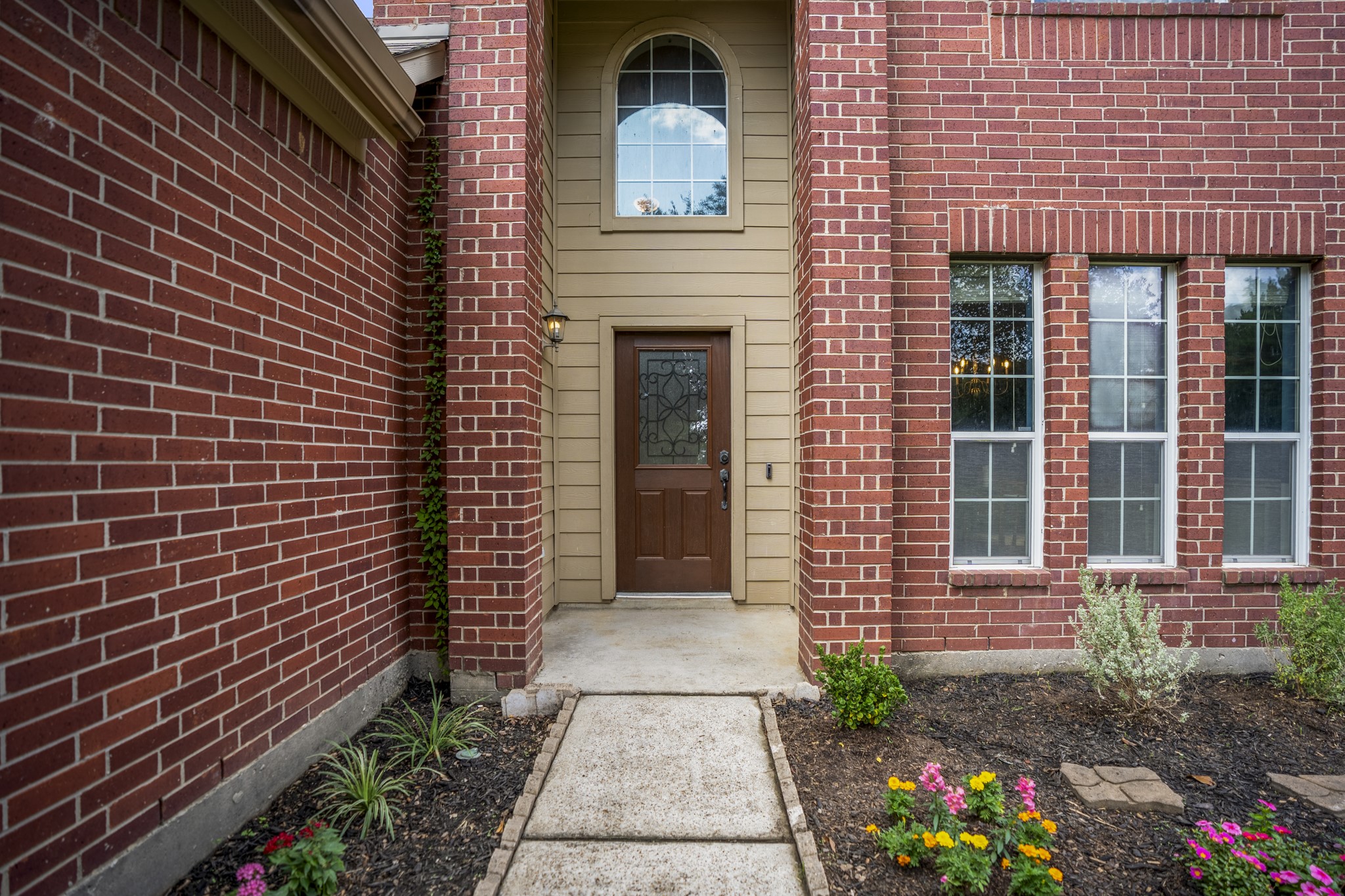 Two-story brick arch detail gives the covered front porch a strong sense of arrival, while siding surrounds the front door and transom window above. Tall front windows nearby bring extra architectural interest and hint at the natural light waiting inside.