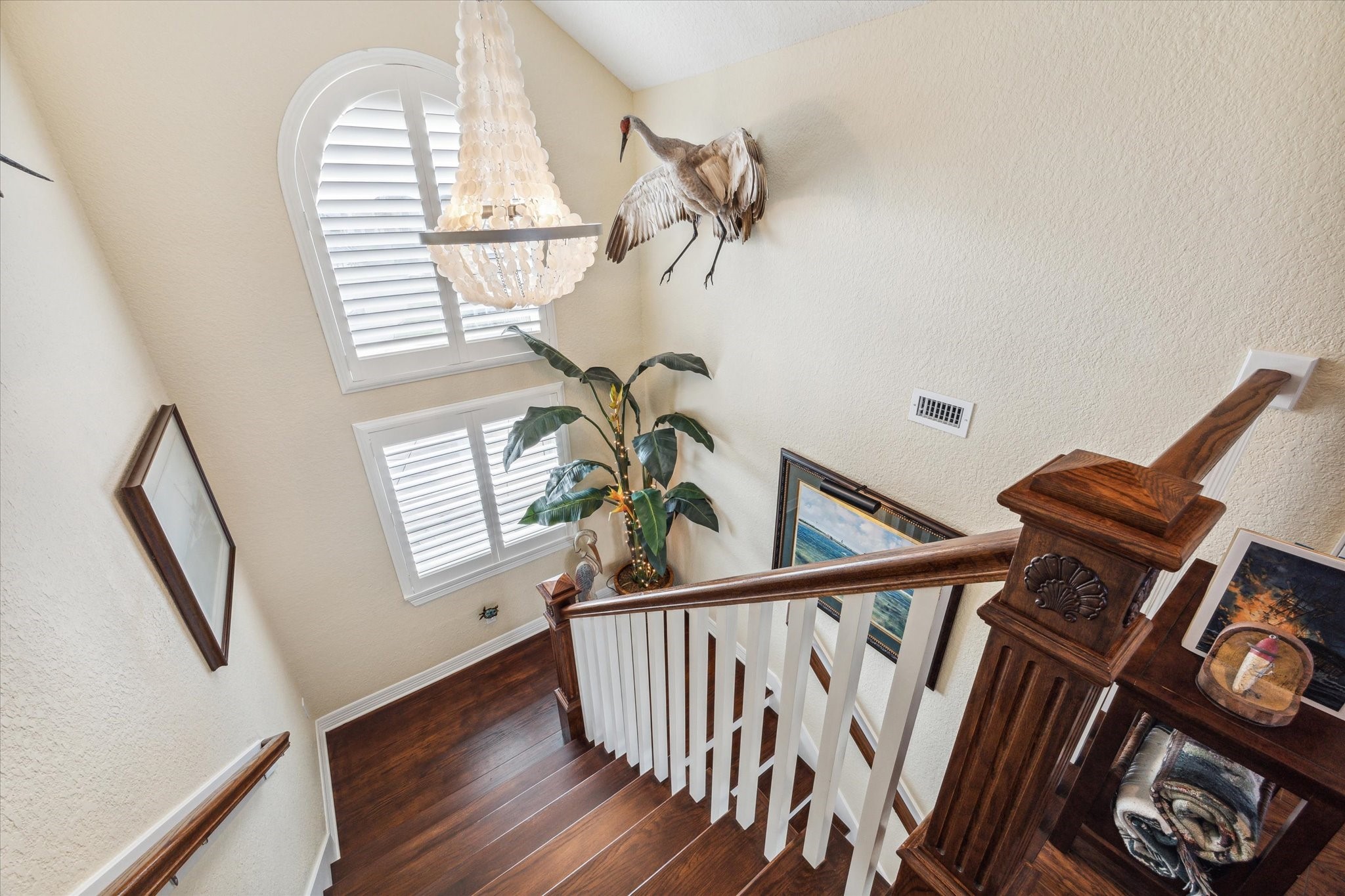 A beautiful arched window with custom plantation shutters sits at the landing going upstairs to the  flex room. A beautiful chandelier adds elegance and interest to the home. In this photo you can also see the custom newel post with seashell detail. No detail was missed in this home!