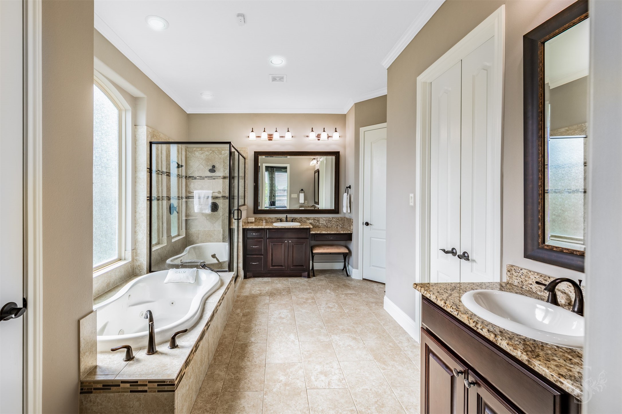 Separate granite-topped vanities with richly stained cabinetry bring elegance and convenience to the spacious primary bath. A jetted tub beneath privacy windows and a separate glass-enclosed shower create an inviting retreat for unwinding.