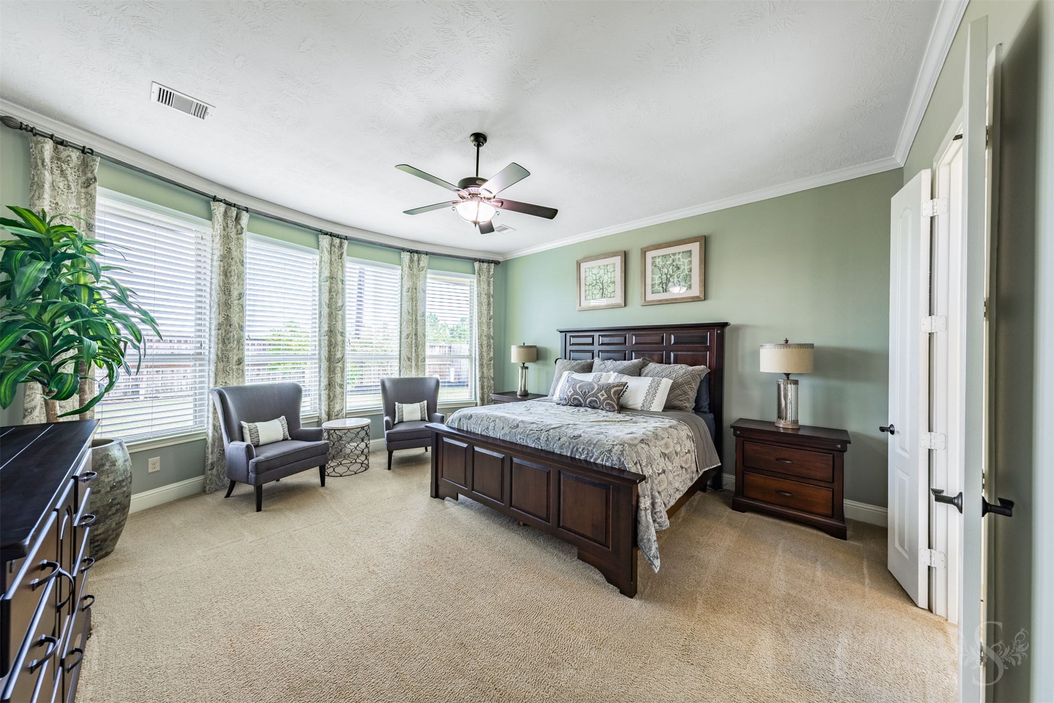 The spacious primary bedroom offers a peaceful retreat with soft green walls, plush carpeting and elegant crown molding. A wall of tall windows brightens the room and provides a lovely backdrop for a comfortable sitting area.