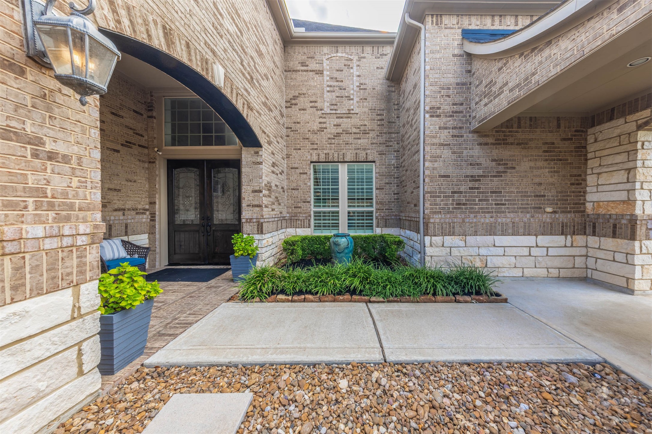 An impressive brick arch frames the covered entrance, where richly stained double doors with decorative glass create an elegant welcome. Stone accents and lush landscaping add beautiful texture to the courtyard setting.