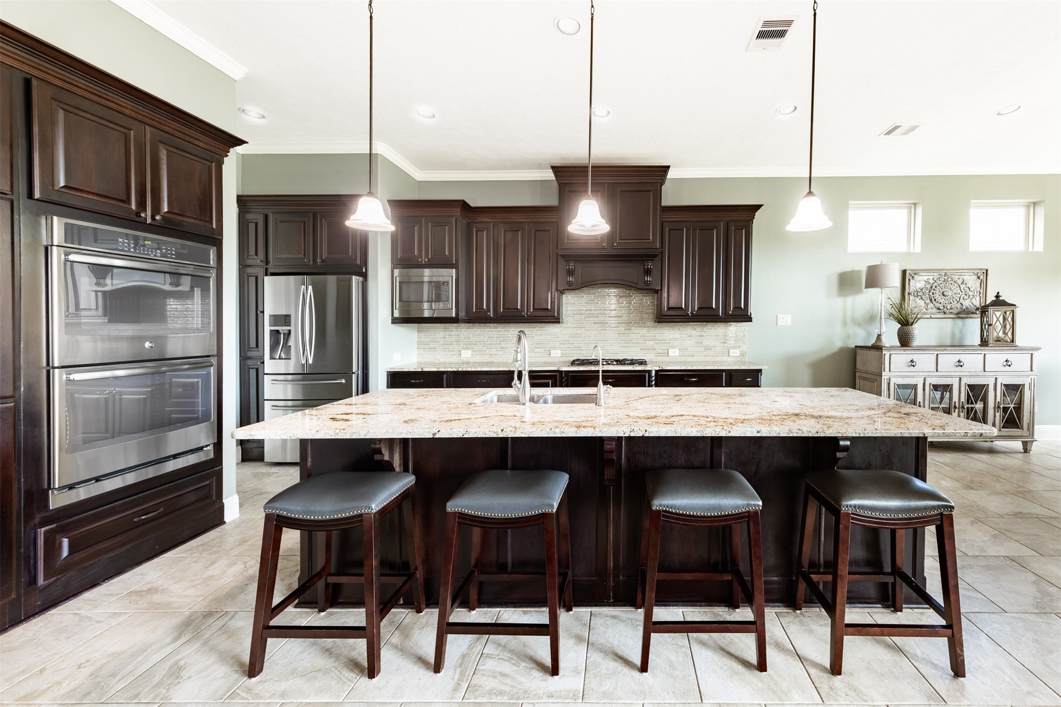 An expansive granite island with generous seating and a trio of pendant lights anchors this beautifully appointed kitchen. Richly stained cabinetry, stainless steel double ovens and a gas cooktop beneath a decorative wood vent hood combine elegant style with everyday convenience.