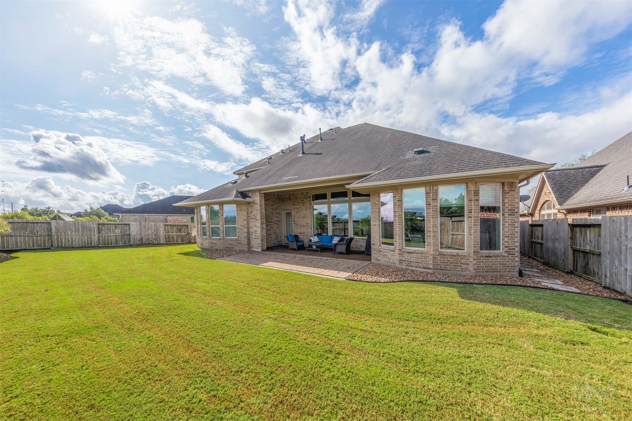 Beautiful bay windows flank the inviting covered patio, creating a striking backdrop for outdoor living. An extended paver patio overlooks the spacious fenced backyard, offering plenty of space for gathering with friends and enjoying time outdoors.