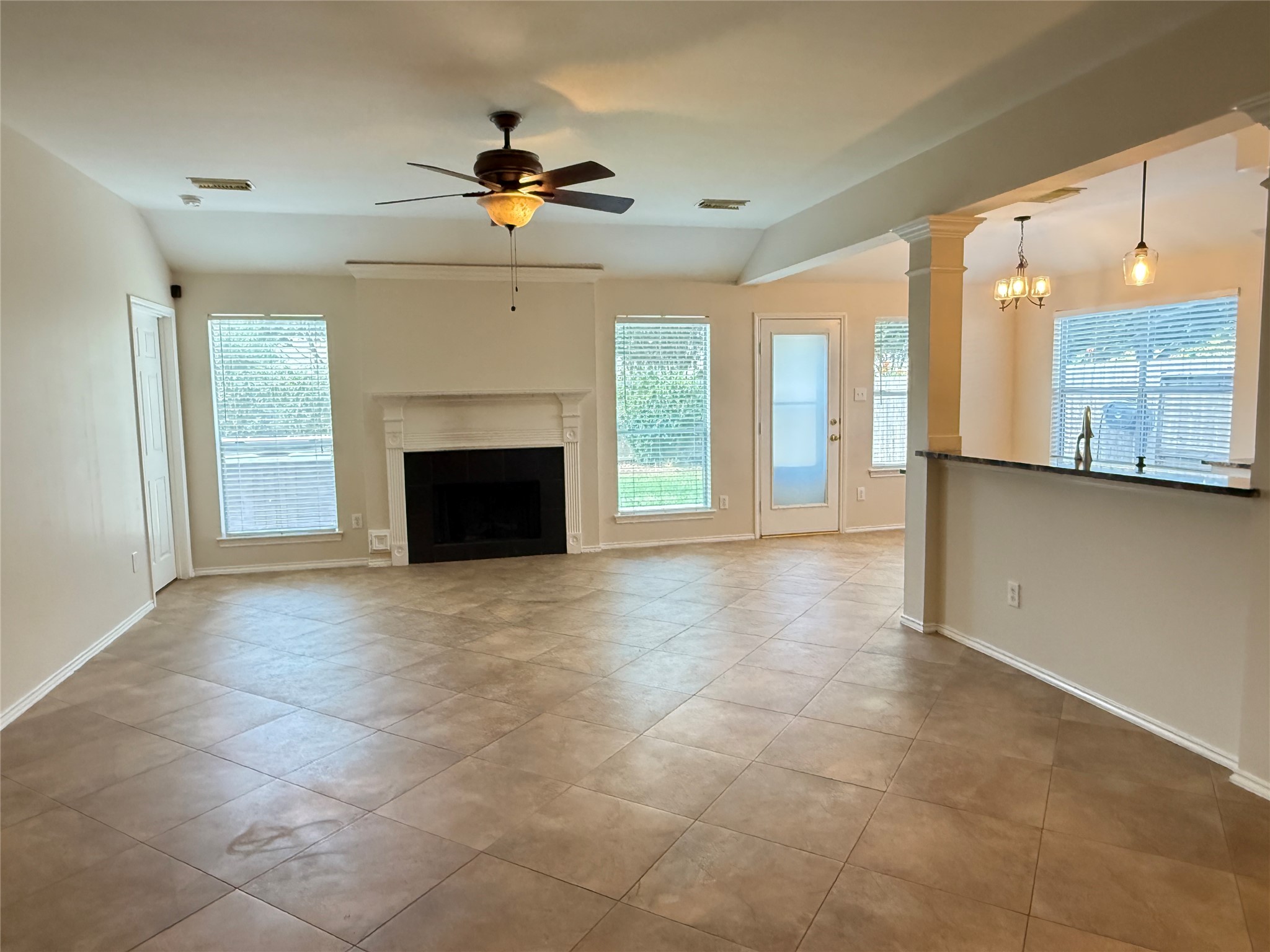 Spacious living area with neutral tile flooring, a cozy fireplace, and abundant natural light from large windows. The open layout seamlessly connects to a modern kitchen, perfect for entertaining.