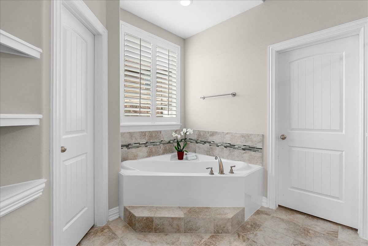 Elegant primary bathroom featuring a luxurious corner soaking tub with stylish tile accents, complemented by natural light streaming through plantation shutters.