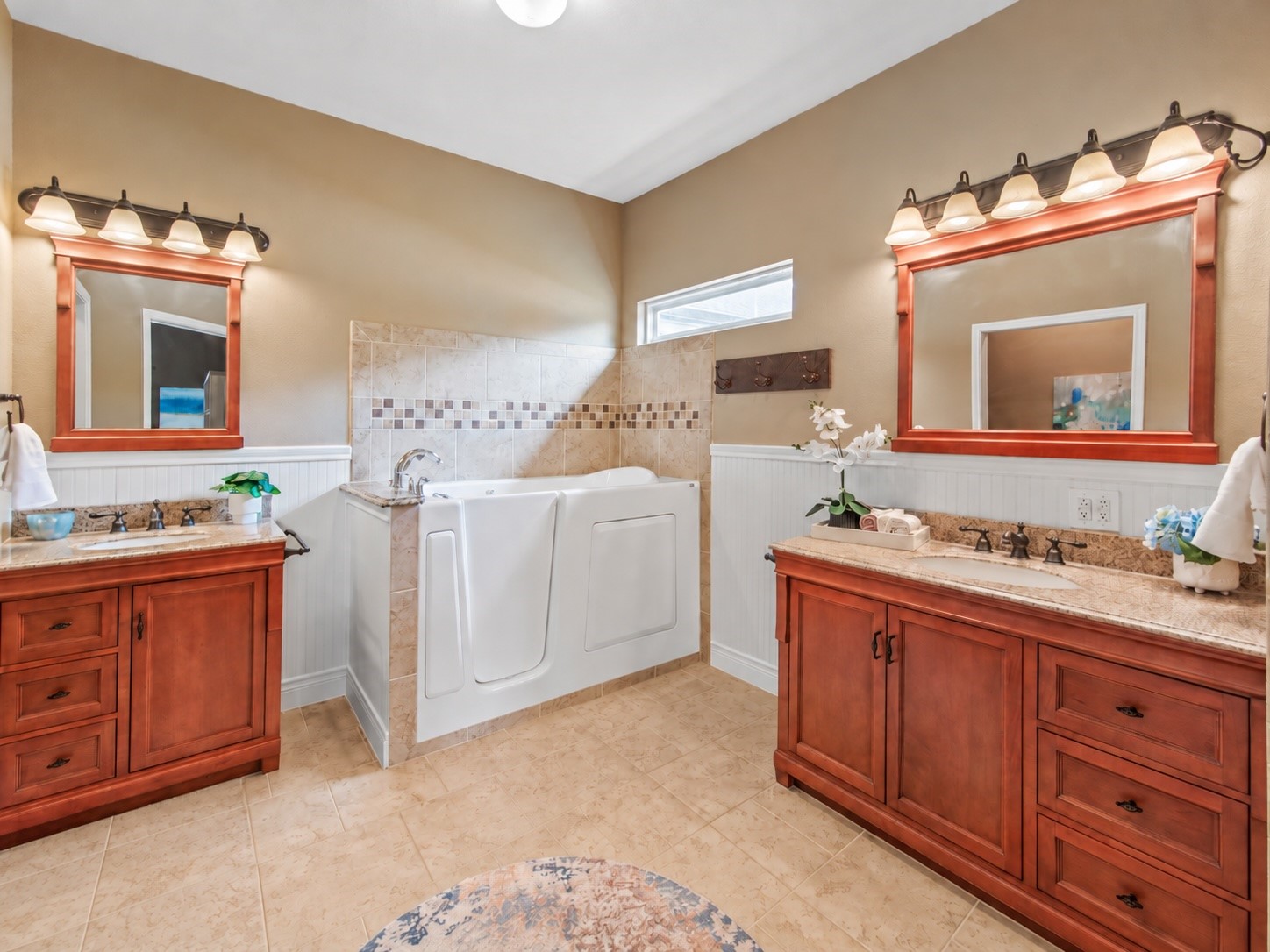 Primary bath featuring two separate vanities with generous storage, a walk-in tub, and decorative tile accents complemented by beadboard wainscoting.