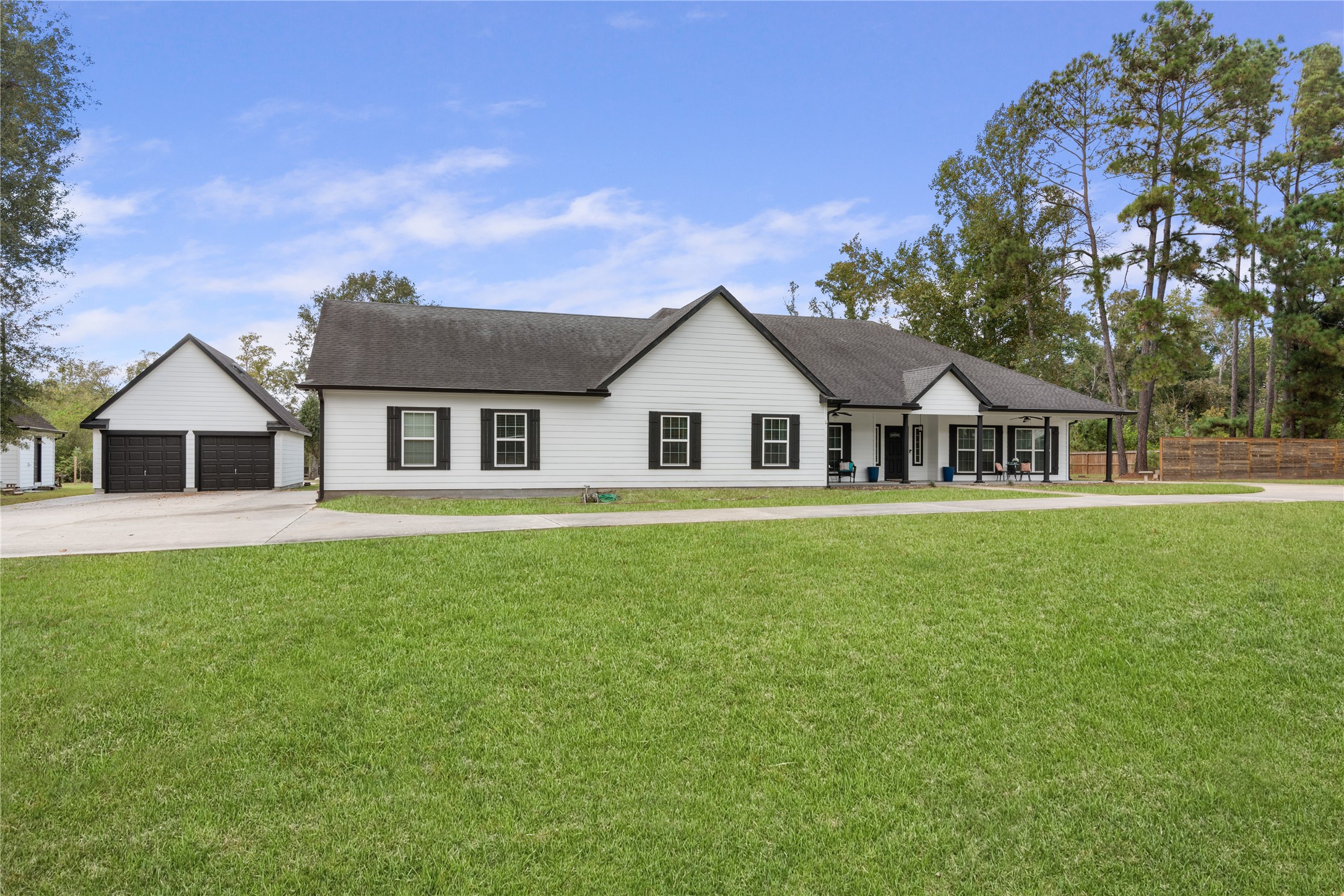 Inviting single-story home featuring a spacious covered front porch, classic white exterior with black accents, detached two-car garage, and an expansive front lawn framed by mature trees.