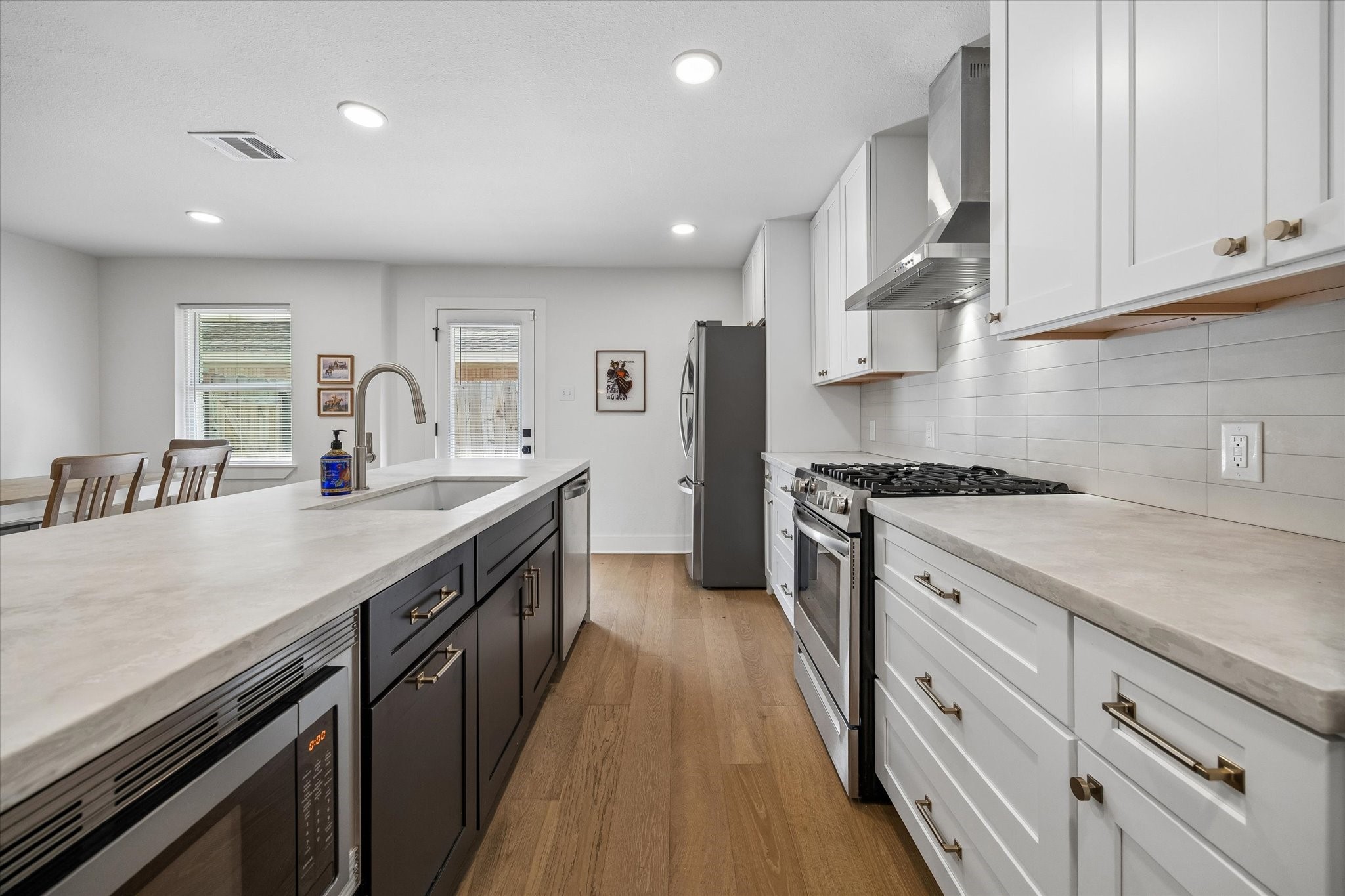 Crisp white cabinetry is balanced by a contrasting island, warm wood flooring and brushed gold hardware. Stainless appliances, a gas range and streamlined vent hood complete the polished kitchen.
