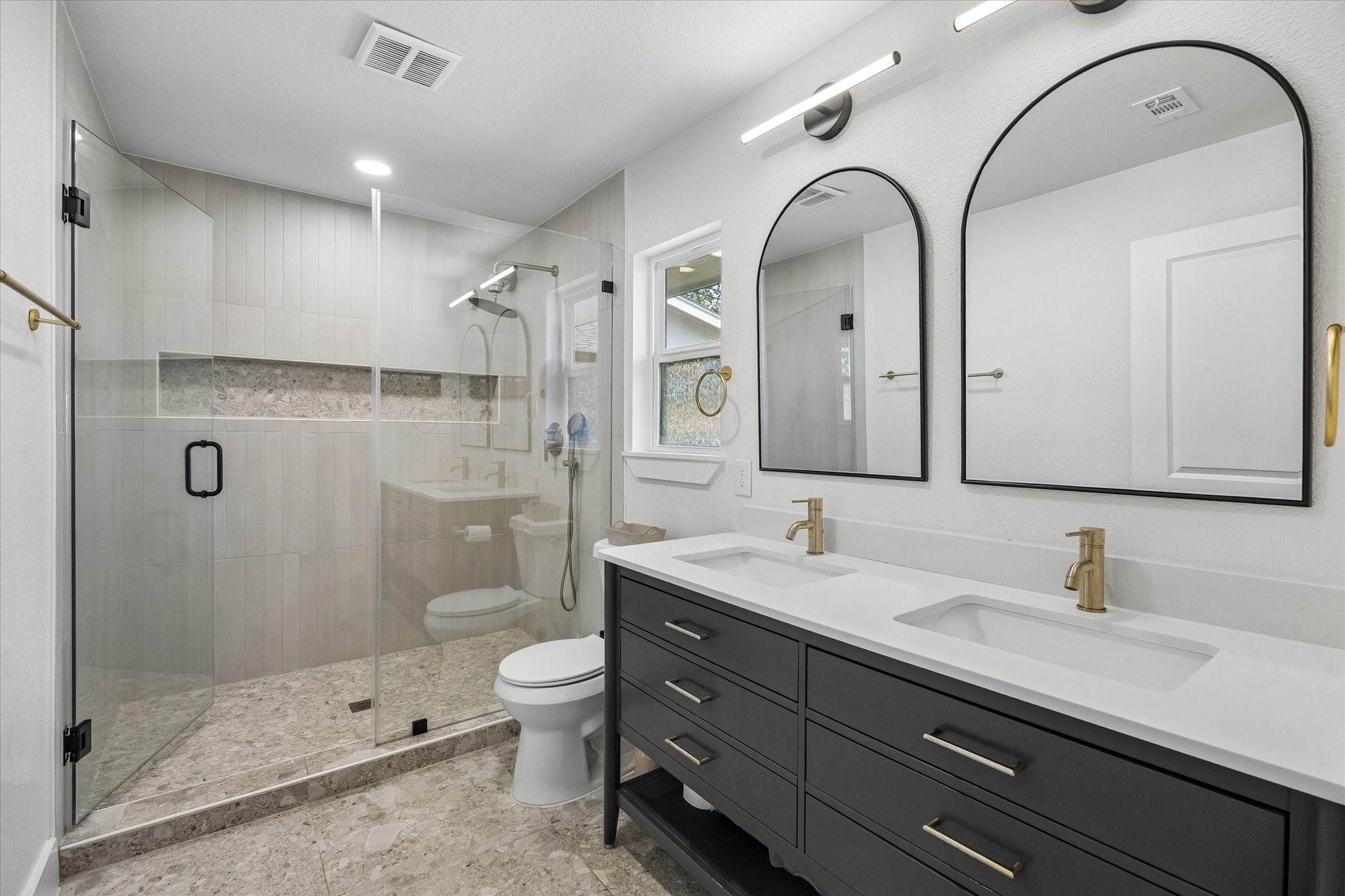 Clean lines give the primary bath a polished, modern feel, pairing a double vanity with warm brass accents and arched mirrors. An oversized glass enclosed shower features floor to ceiling tile, a built in niche, and handheld shower fixture.
