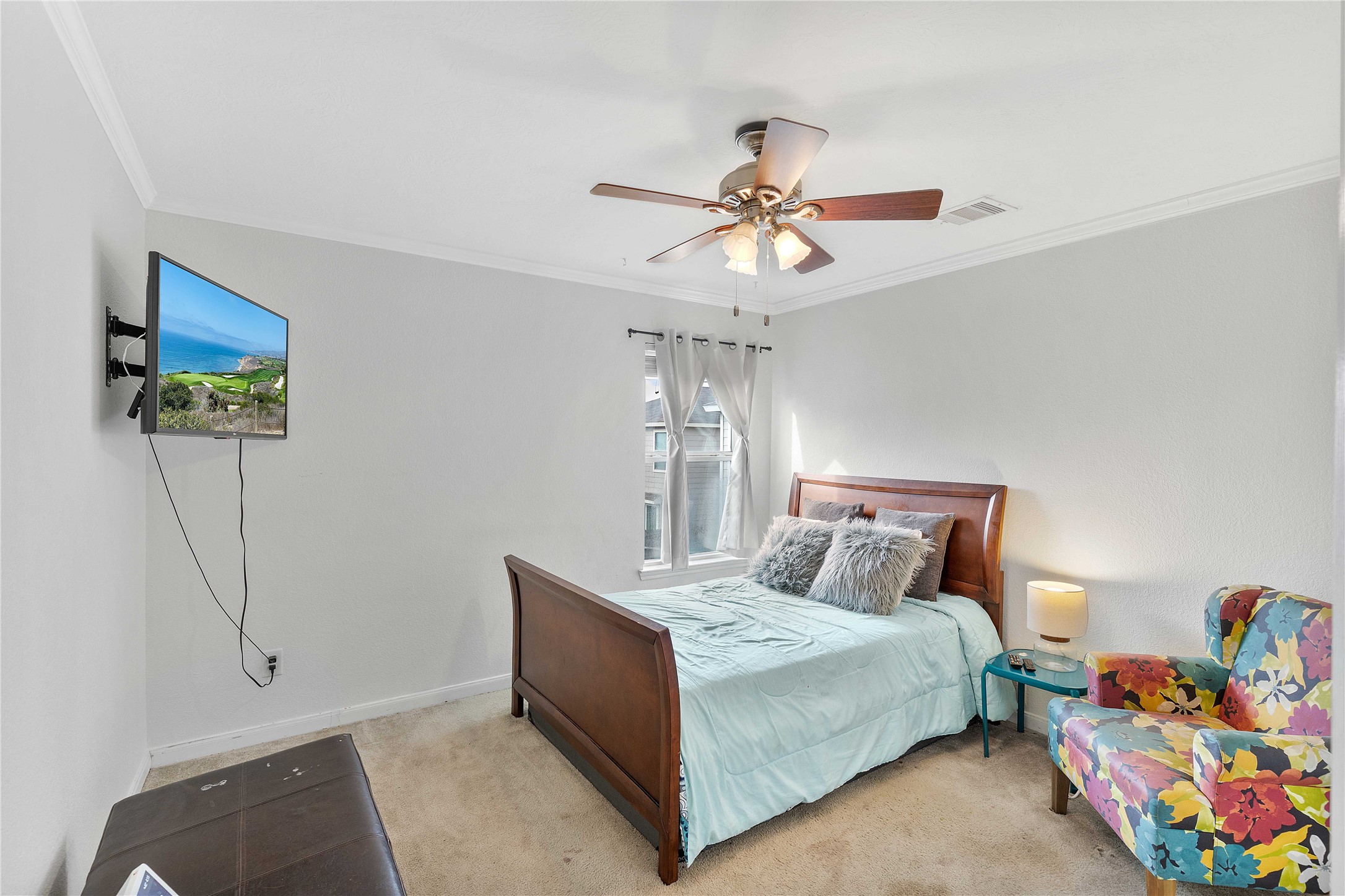 Cozy bedroom with a stylish ceiling fan, wall-mounted TV, and ample natural light. Features a comfortable bed with plush pillows and a colorful accent chair for added charm.
