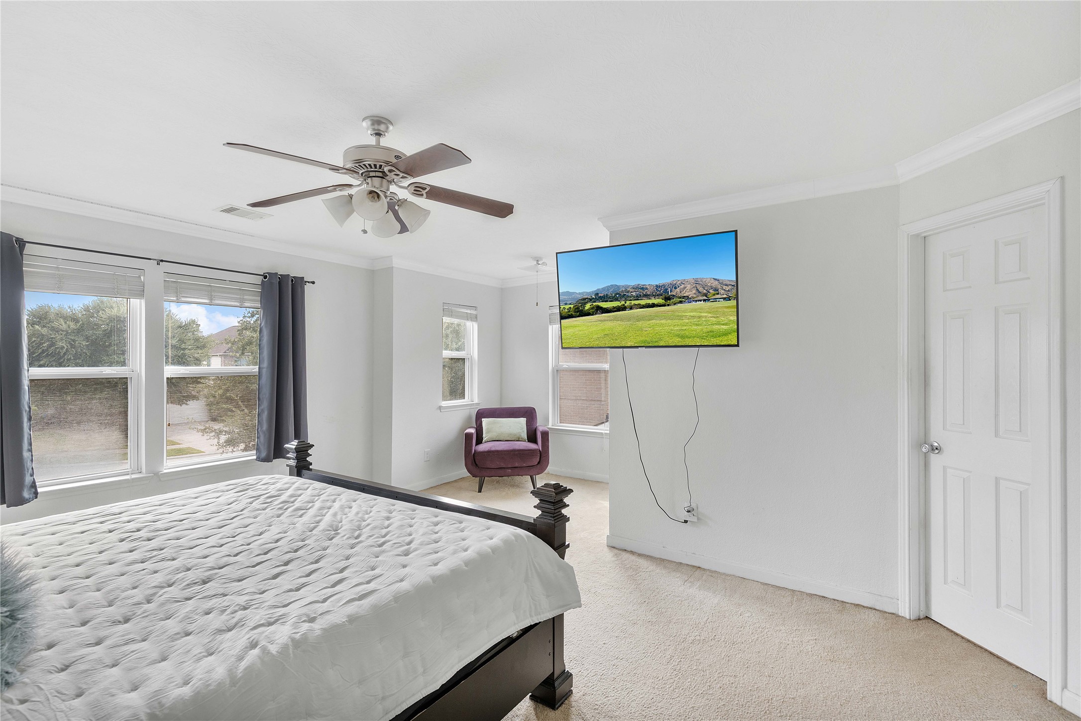 Spacious and bright bedroom with large windows, modern ceiling fan, and a wall-mounted TV, offering a cozy and inviting ambiance.
