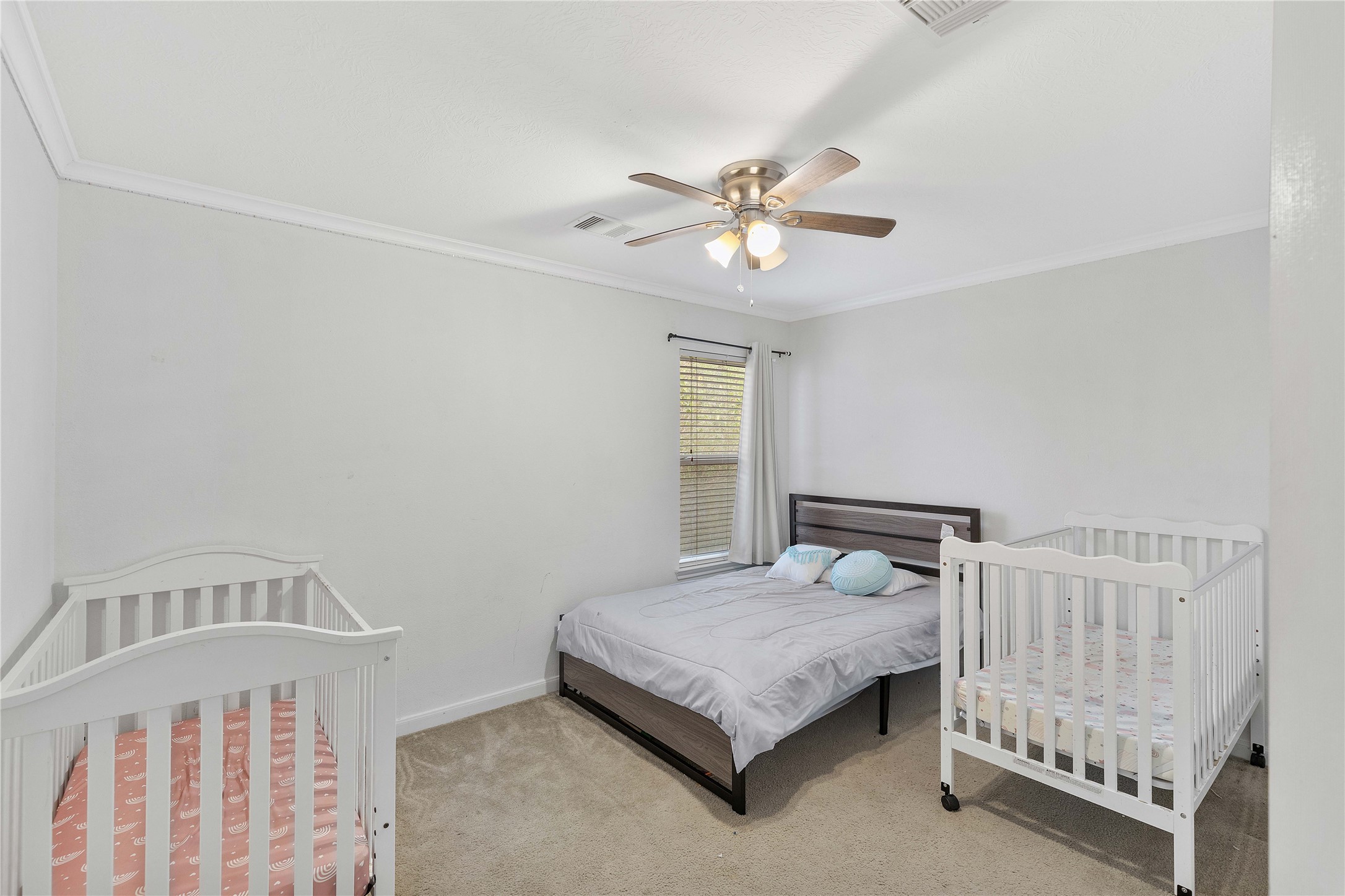 Charming bedroom with a cozy layout, featuring a comfortable bed, two cribs, and a ceiling fan, all bathed in natural light from a large window.