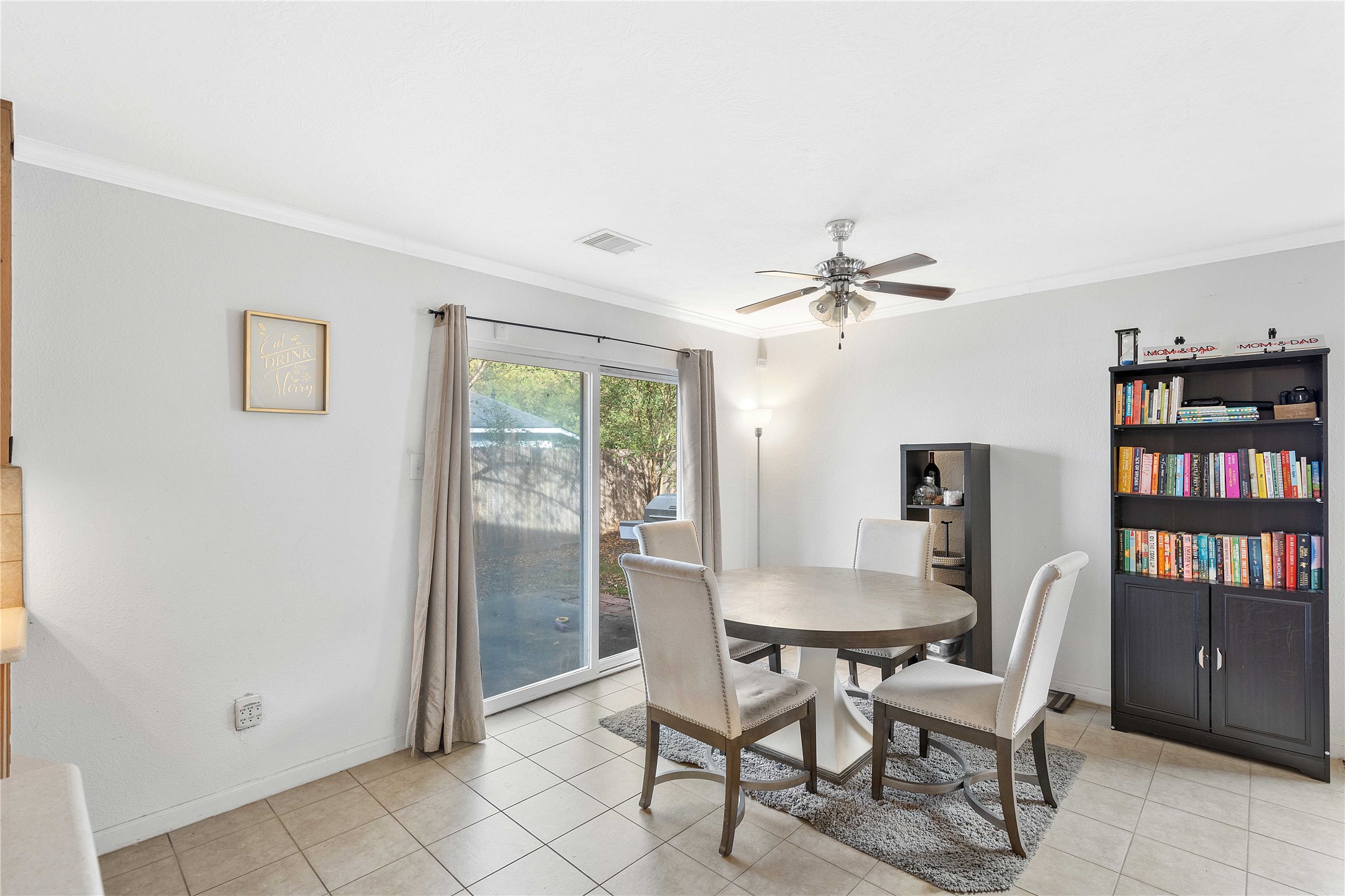 Bright dining area with tile flooring features a round table, comfortable chairs, and a ceiling fan. A large sliding glass door offers access to a serene backyard, while a bookshelf adds charm and functionality.