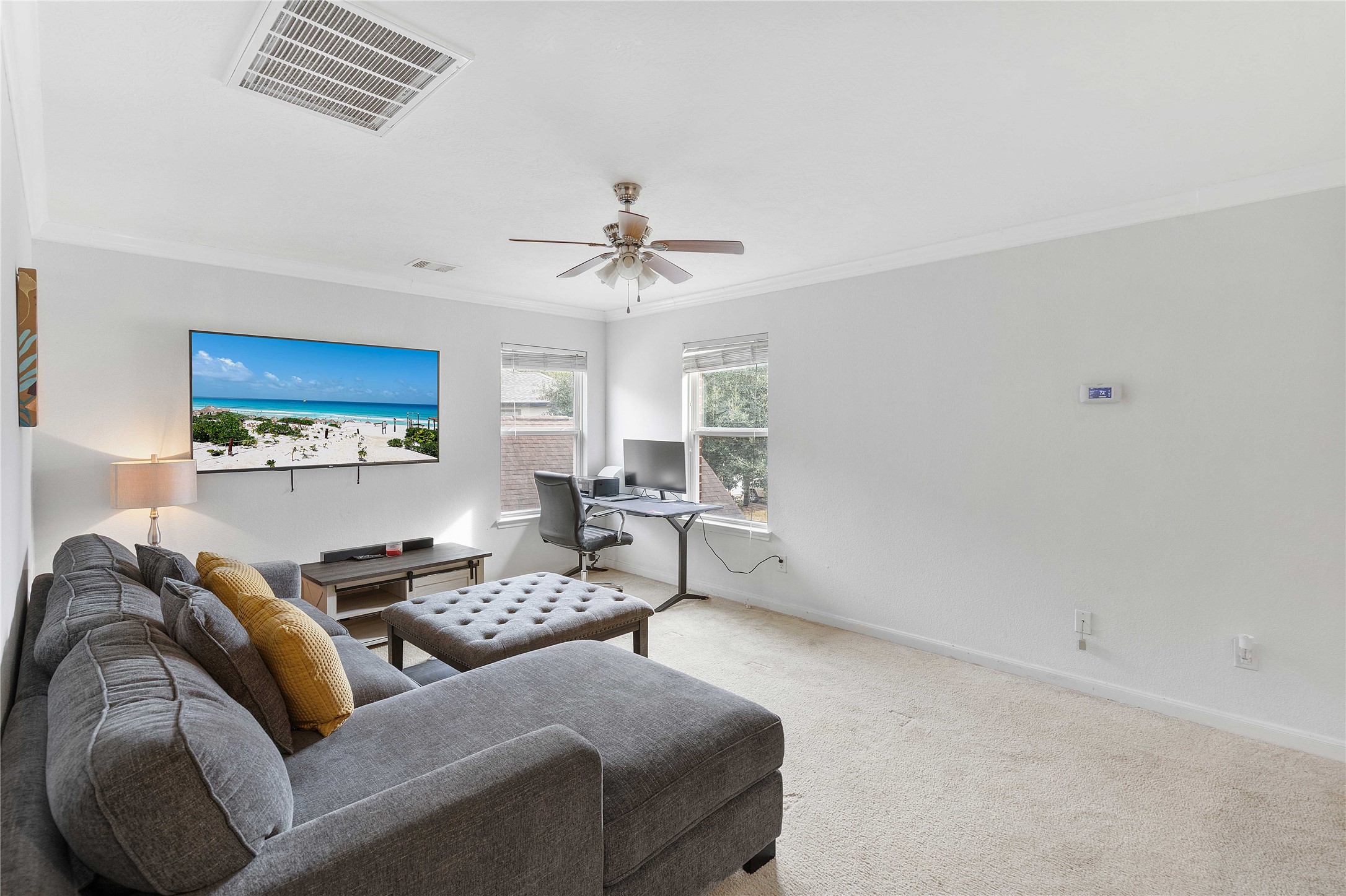 Spacious and bright living area with a cozy sectional sofa, wall-mounted TV, and a convenient corner office desk, perfect for relaxation and productivity.