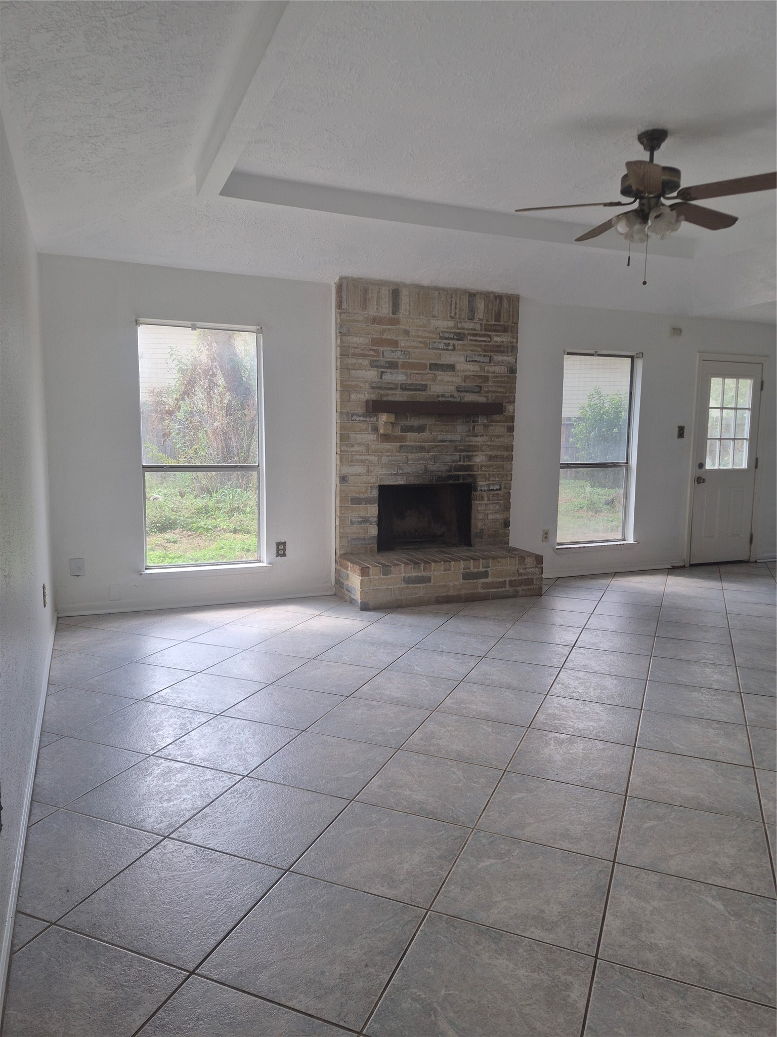 Spacious living area with a brick fireplace, tile flooring, and natural light from two large windows.