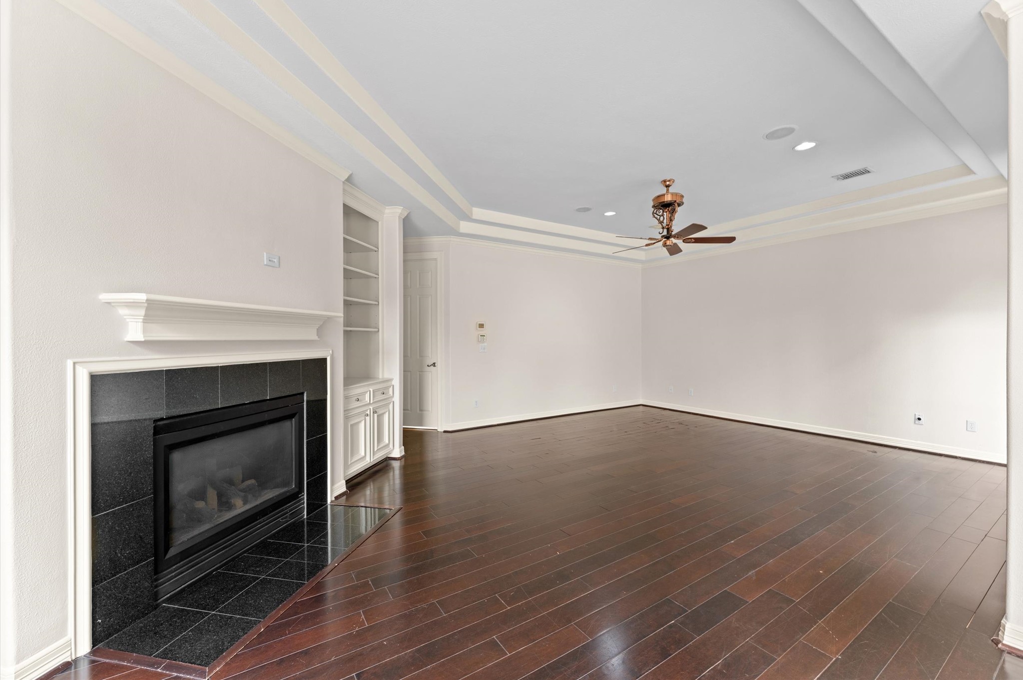This view of the primary bedroom, highlights a gas fireplace with mantle, built-in bookcase, wood flooring and tray ceiling.