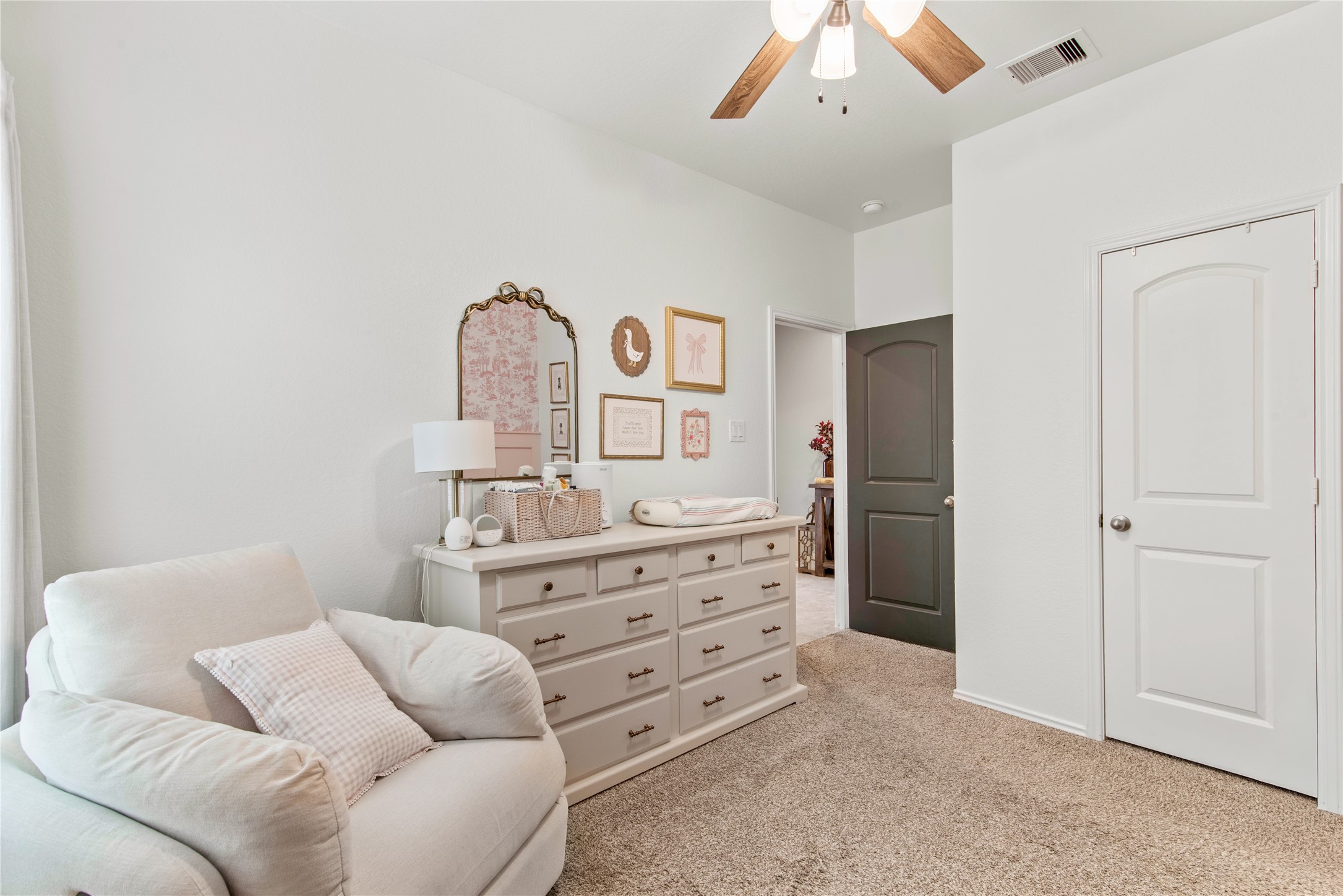 Charming nursery with soft carpet, featuring a cozy armchair and a spacious dresser under a ceiling fan, creating a serene and inviting space.