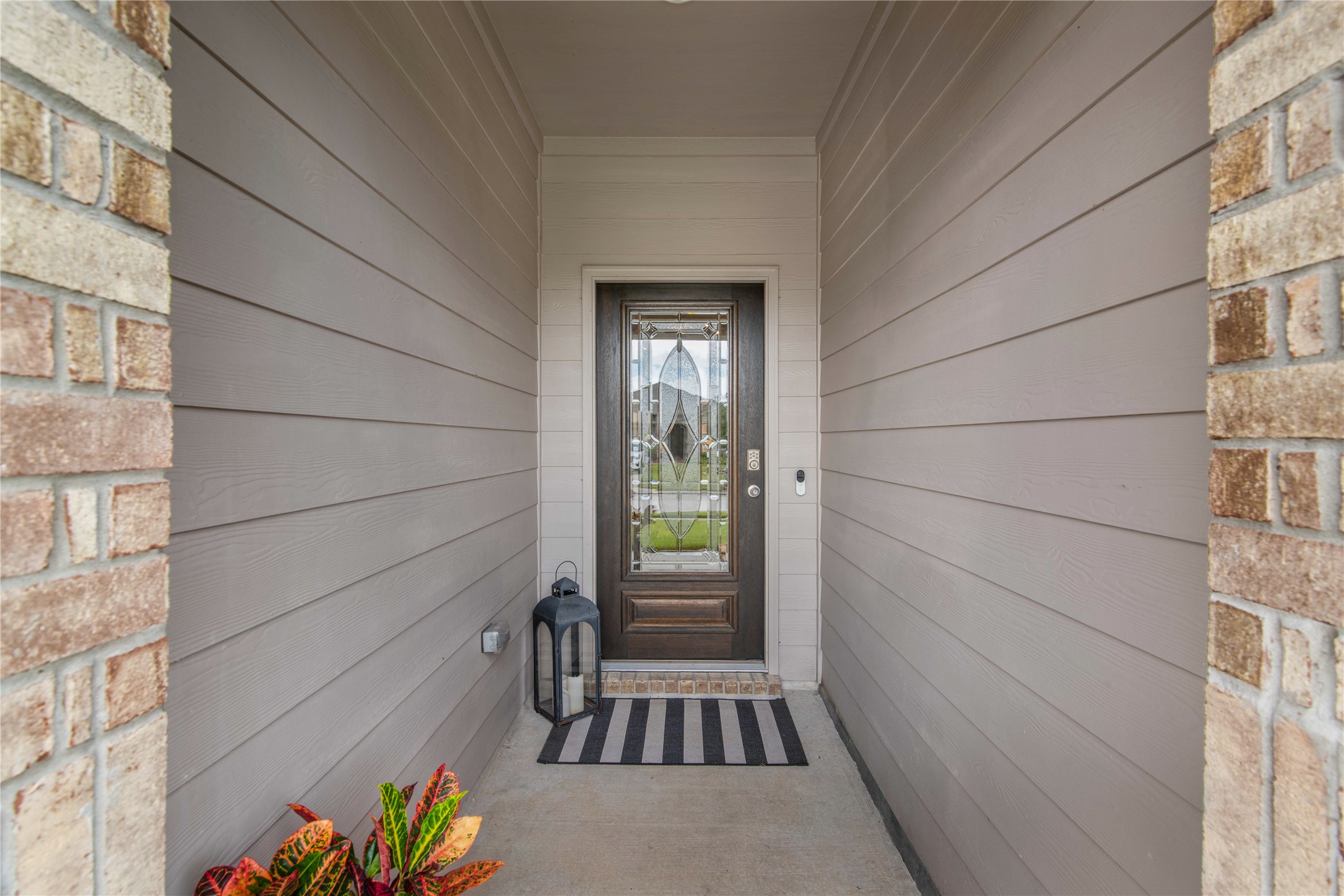 Inviting entryway features a stylish glass-paneled door, sheltered by elegant siding, and accented with a charming lantern and vibrant potted plant.