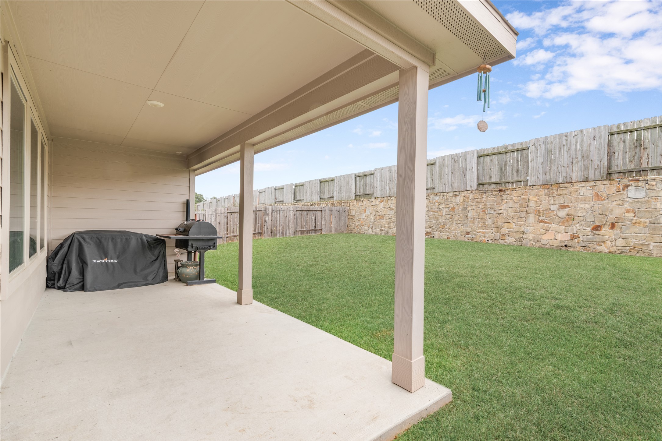Spacious covered patio overlooking a lush, fenced backyard with a charming stone wall, perfect for outdoor entertaining and relaxation.