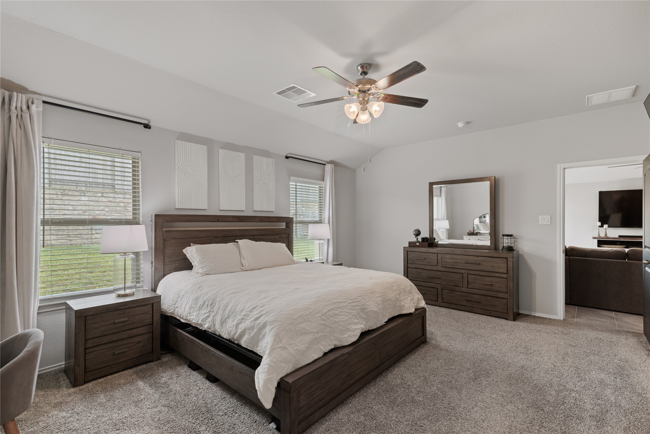 Spacious and inviting master bedroom featuring a cozy carpeted floor, elegant ceiling fan, and ample natural light from dual windows. The room includes a stylish wood bed frame, matching nightstands, and a dresser with a large mirror, exuding comfort and modern charm.