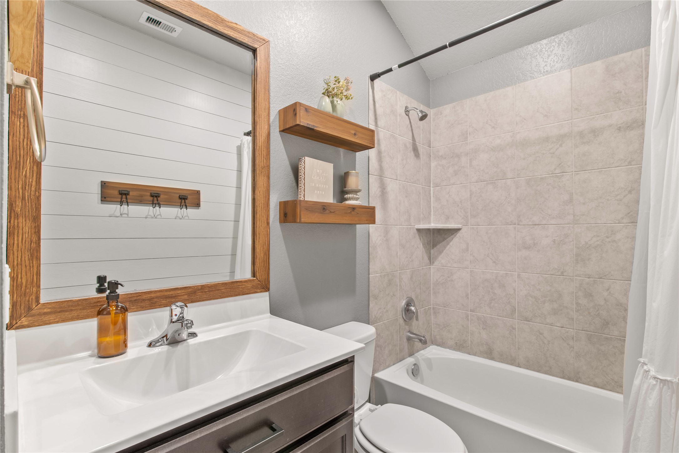 Charming bathroom featuring a sleek vanity with a wooden framed mirror, elegant shiplap wall, and a cozy bathtub with stylish tile surround.