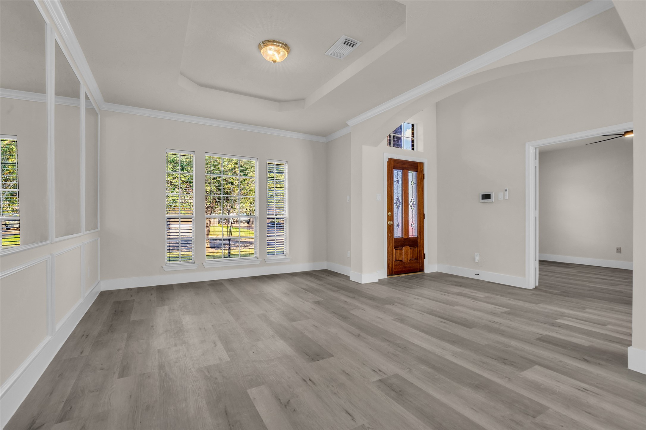 Bright and inviting entryway featuring elegant tray ceilings, large windows for natural light, and sleek wood flooring, complemented by a classic wooden front door.