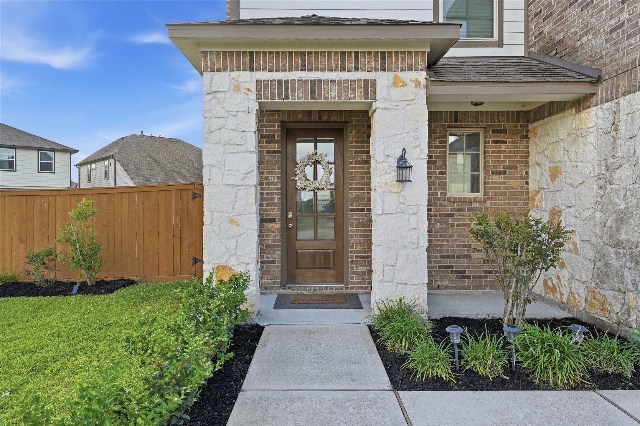 Inviting front entrance with a mix of brick and stone exterior. Wooden door and the pathway is bordered by neat landscaping. A wooden fence encloses the green lawn, adding privacy and charm. Inviting front entrance with a mix of brick and stone exterior. Wooden door and the pathway is bordered by neat landscaping. A wooden fence encloses the green lawn, adding privacy and charm.