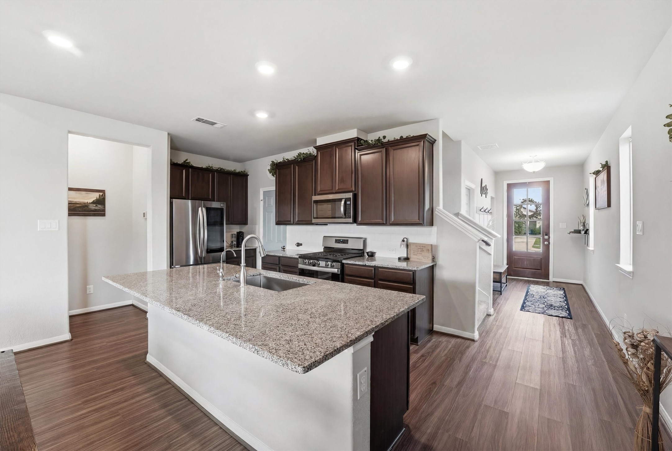 This modern kitchen includes a large island with a sink, stainless steel appliances, and dark wood cabinets. The open layout leads to an entryway with a wooden floor and a glass-paneled front door, creating a bright and inviting space. This modern kitchen includes a large island with a sink, stainless steel appliances, and dark wood cabinets. The open layout leads to an entryway with a wooden floor and a glass-paneled front door, creating a bright and inviting space.