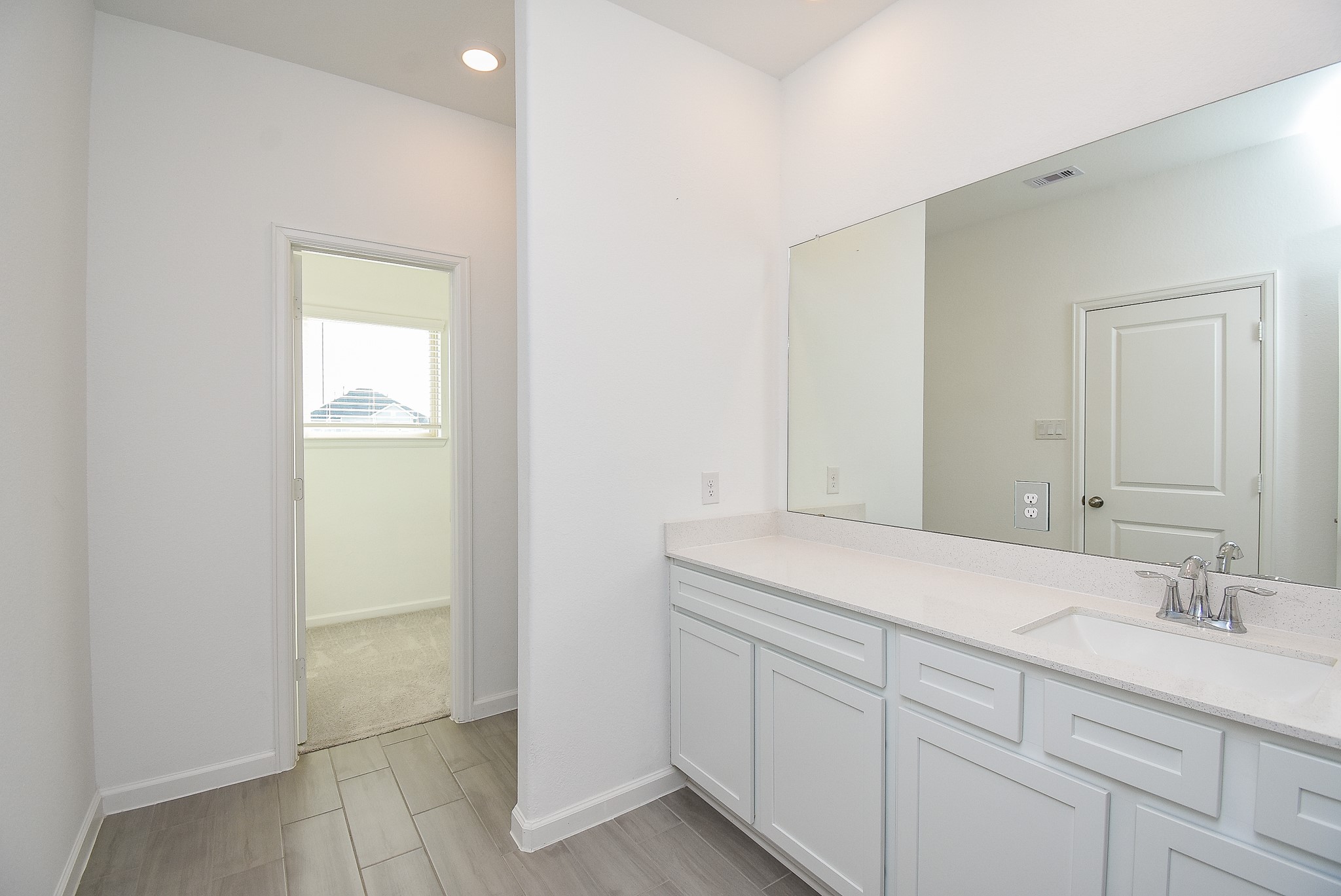 A bright and spacious primary bathroom featuring a large vanity with dual sinks, white cabinetry, and a wide mirror. The light tiled flooring and recessed lighting enhance the modern, airy feel. The walk-in closet is visible through the open doorway, providing convenient storage.