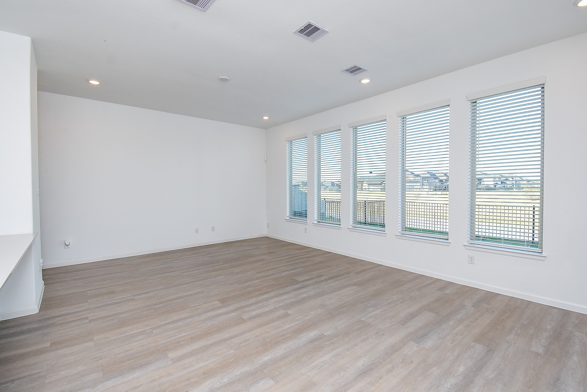 A spacious and bright living area featuring large windows with blinds, allowing plenty of natural light and offering a scenic outdoor water view. The light wood flooring enhances the open feel, while the recessed lighting and neutral white walls create a modern and inviting atmosphere.