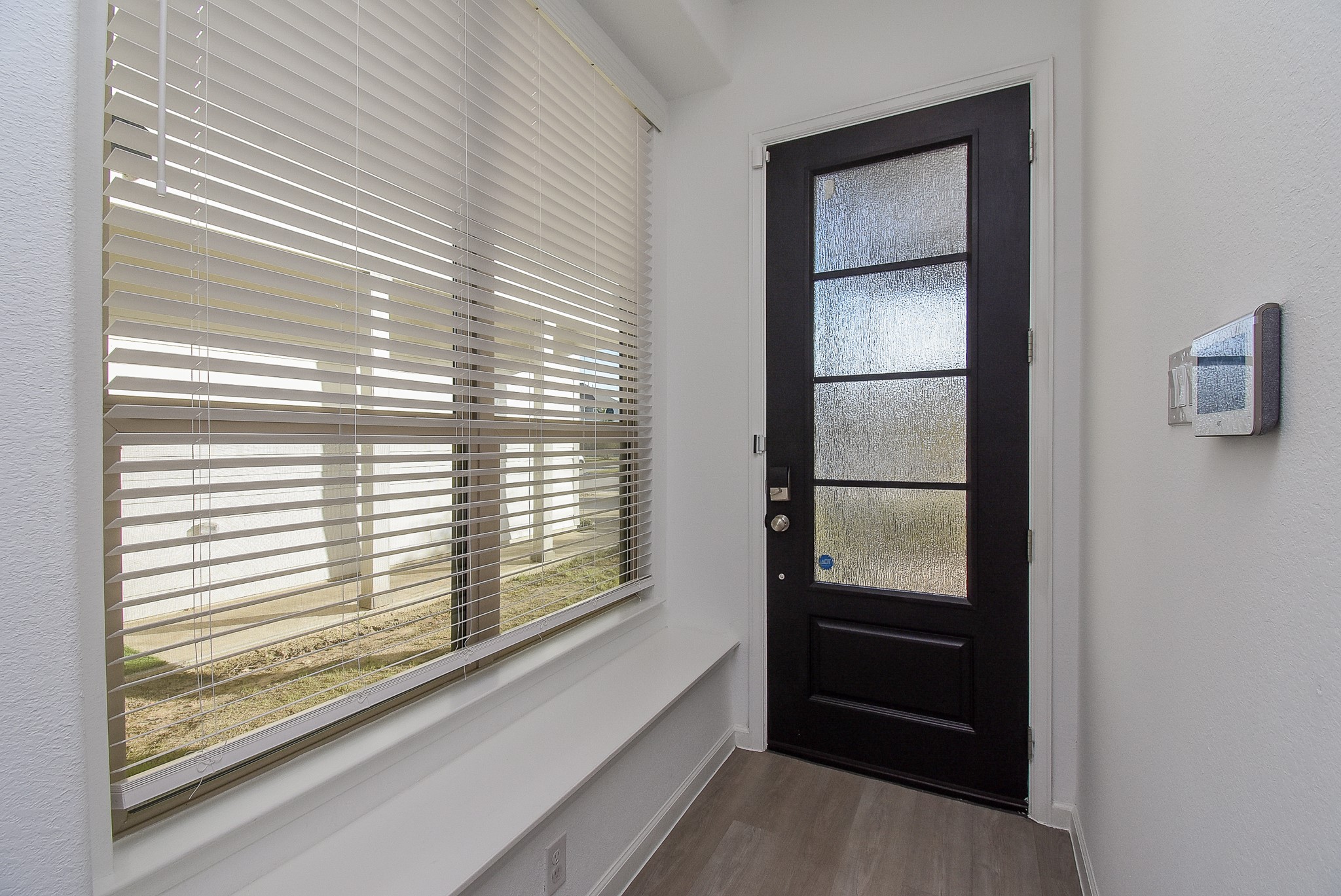 A modern entryway interior featuring a large window with white blinds allowing the natural light to filter in, and a built-in bench runs along the wall