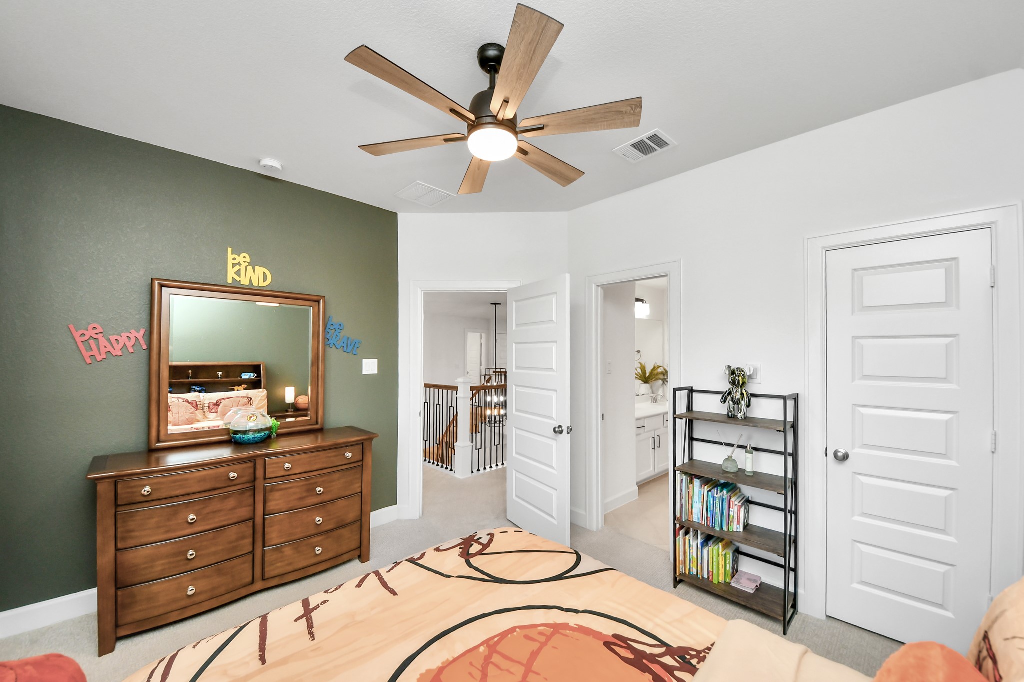 Cozy bedroom with a stylish green accent wall, modern ceiling fan, and ample natural light. Features a wooden dresser with a mirror, open shelving for books and decor, and access to a well-lit bathroom.