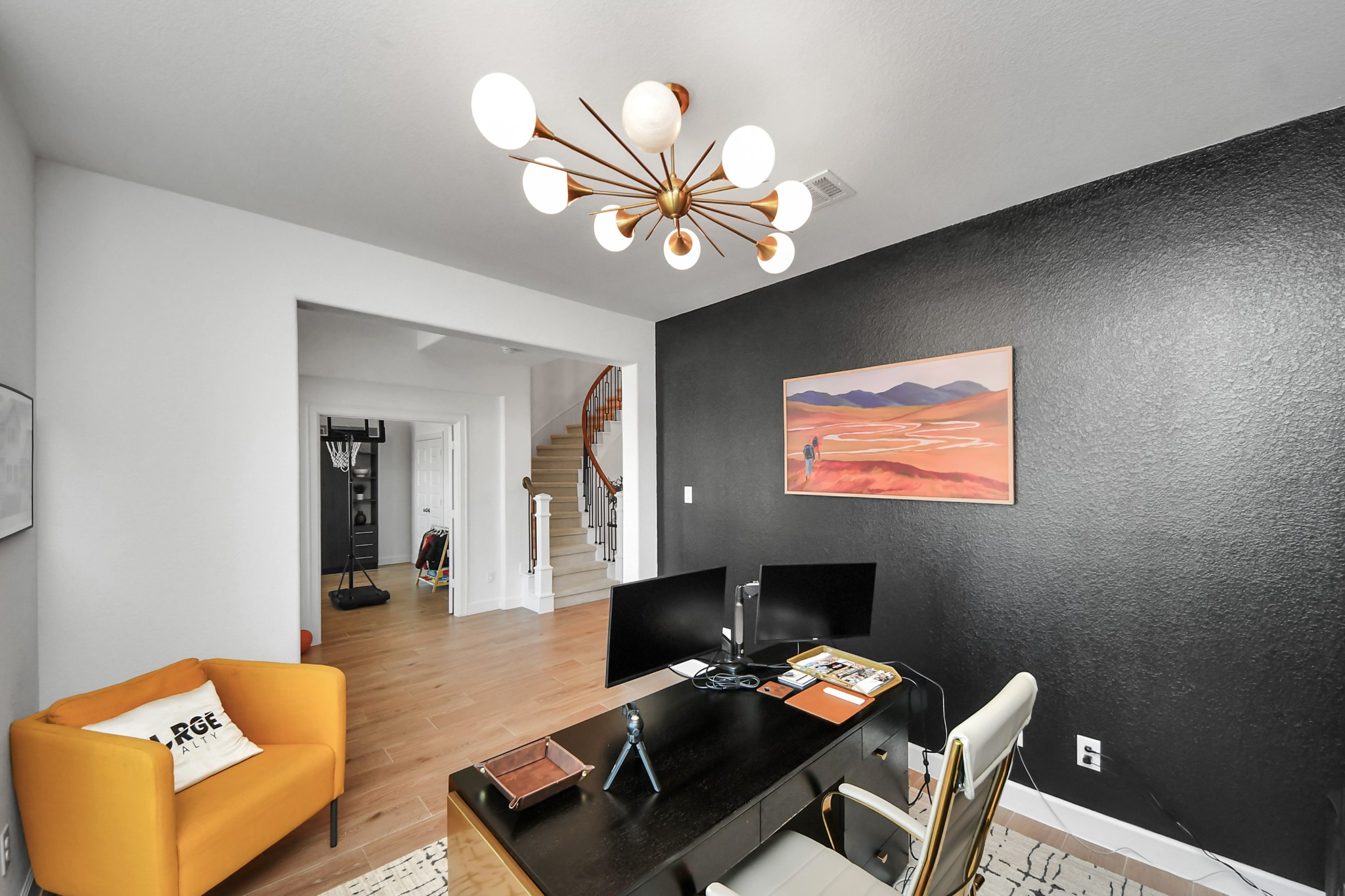 Stylish home office featuring a bold black accent wall, modern light fixture, and warm wood flooring, with an inviting open view to the staircase.