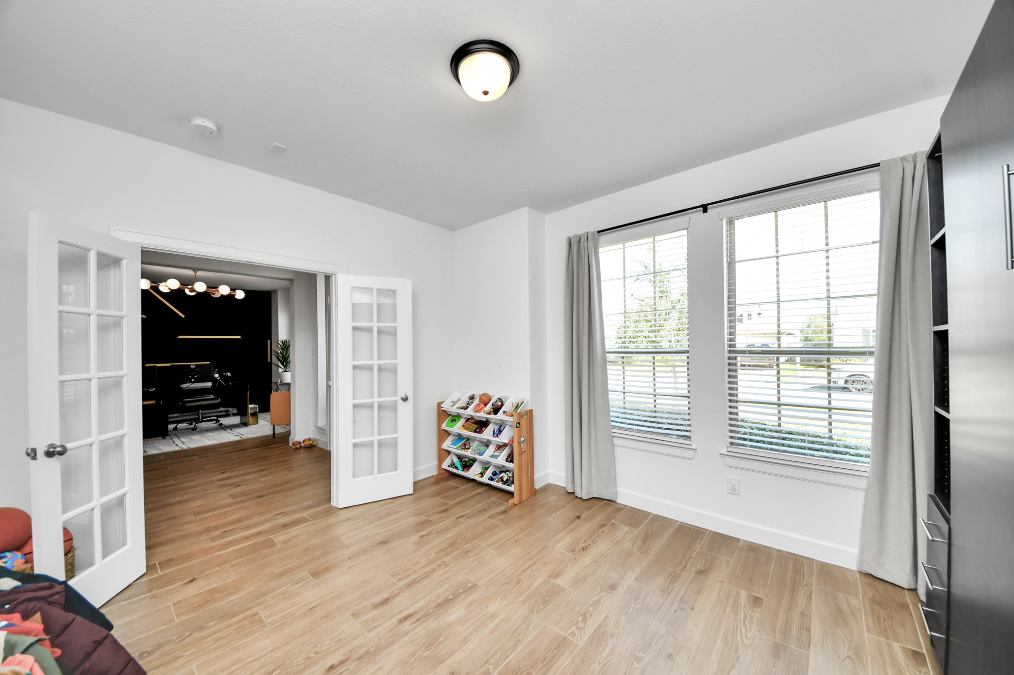 Bright and spacious room with elegant wood flooring, large windows for natural light, and charming French doors leading to an adjacent room, perfect for a home office or play area.