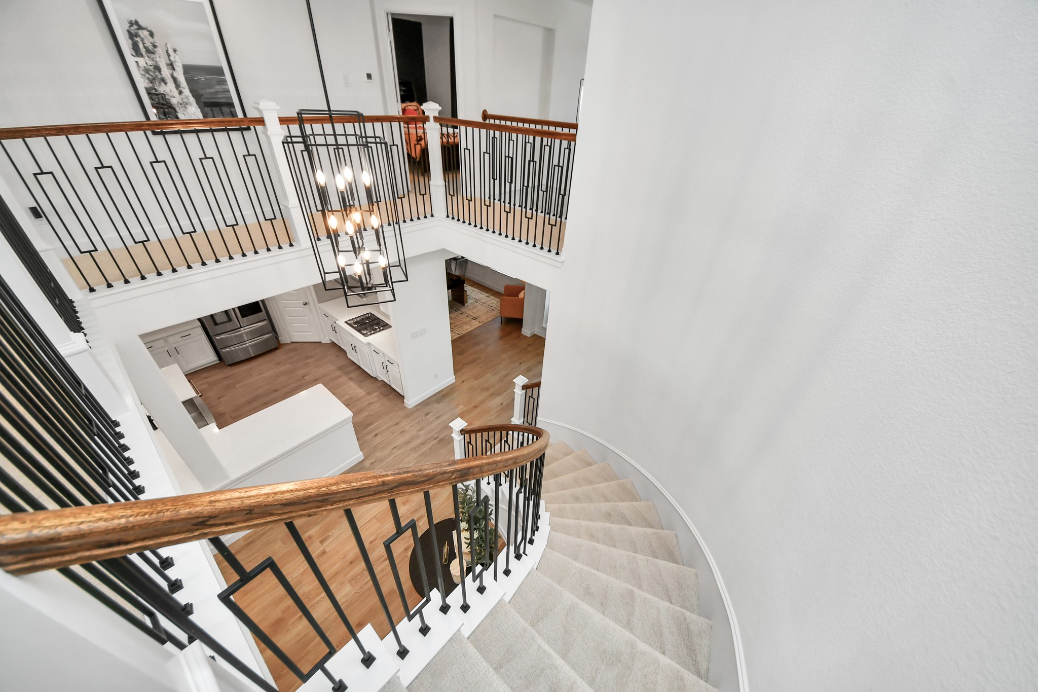 Elegant curved staircase with wrought iron railings leads to an open upper landing, overlooking a modern kitchen with sleek appliances and hardwood floors.