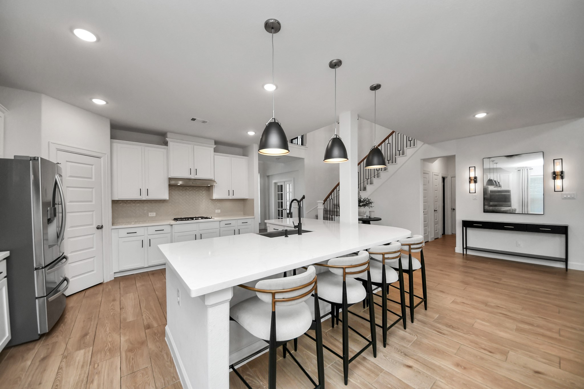 Spacious and modern kitchen with sleek white cabinetry, a large center island with seating, stainless steel appliances, and elegant pendant lighting, seamlessly flowing into an open living area with a stylish staircase.