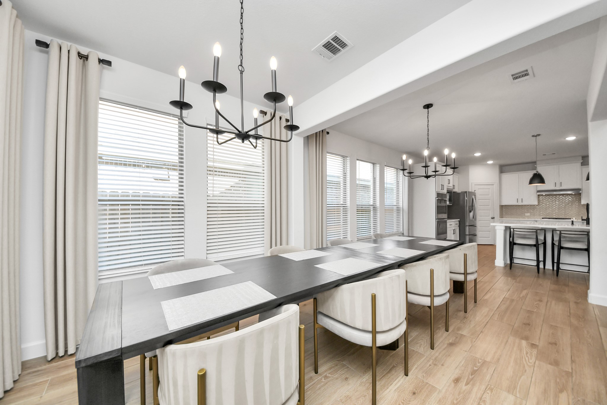 Elegant dining area with a long table, modern chandelier, and large windows offering abundant natural light, seamlessly flowing into a sleek kitchen with stainless steel appliances. Elegant dining area with a long table, modern chandelier, and large windows offering abundant natural light, seamlessly flowing into a sleek kitchen with stainless steel appliances.