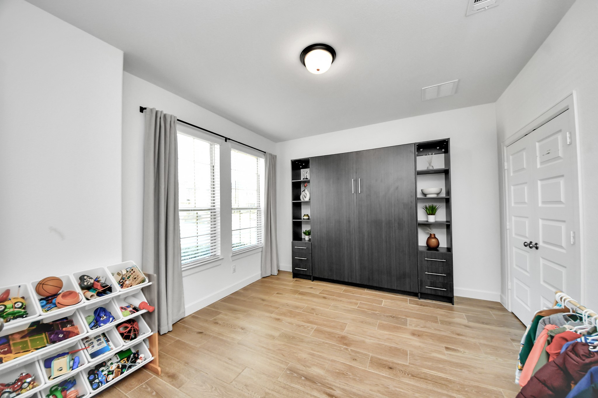 Bright and versatile room featuring a built-in Murphy bed with shelving, large window for natural light, and stylish wood flooring, perfect for a guest room or play area.