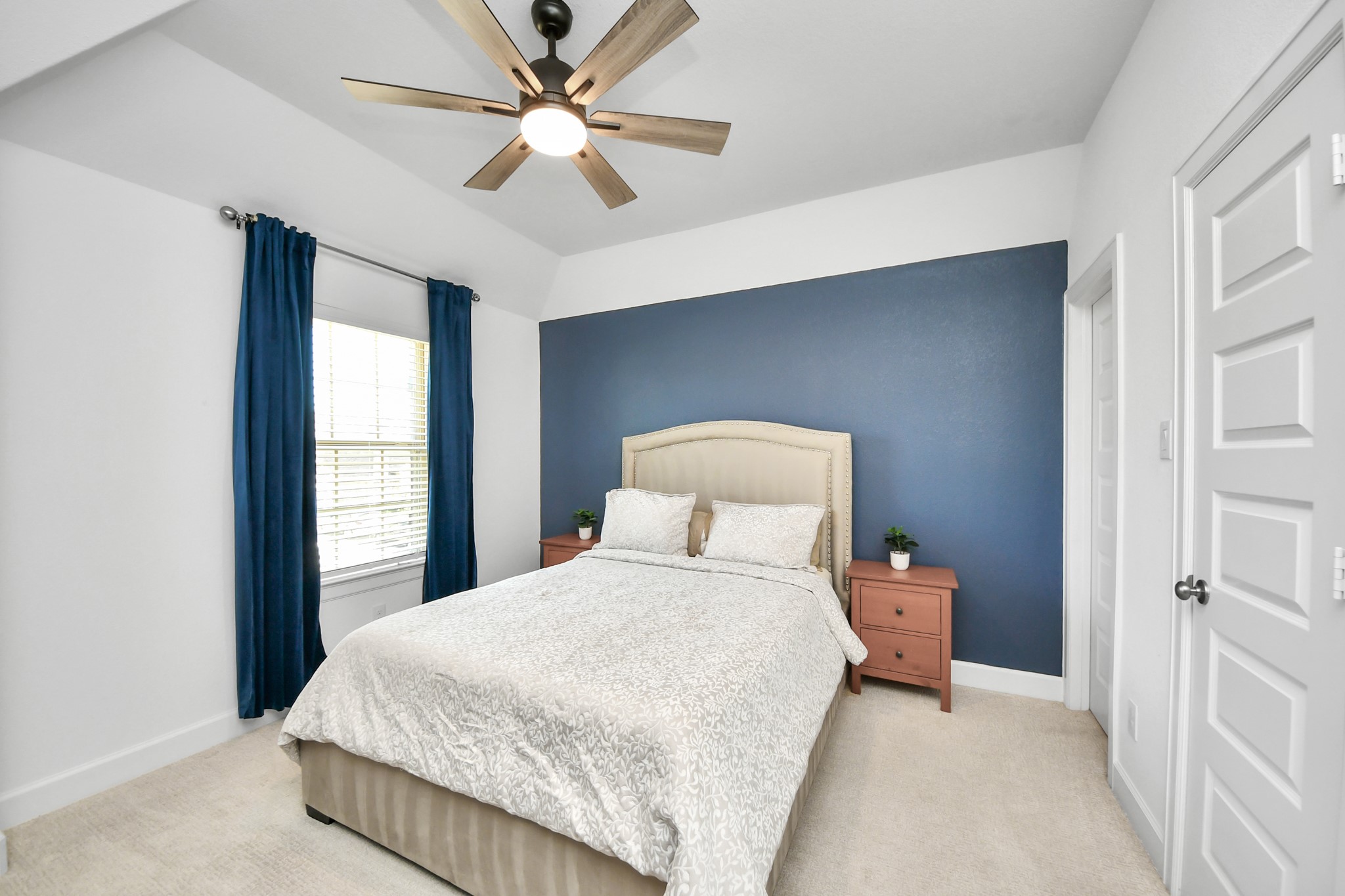 Cozy bedroom featuring a plush bed with a stylish headboard, accented by a bold blue feature wall. Natural light pours in through the window, complemented by elegant blue curtains. A modern ceiling fan enhances the room's comfort.