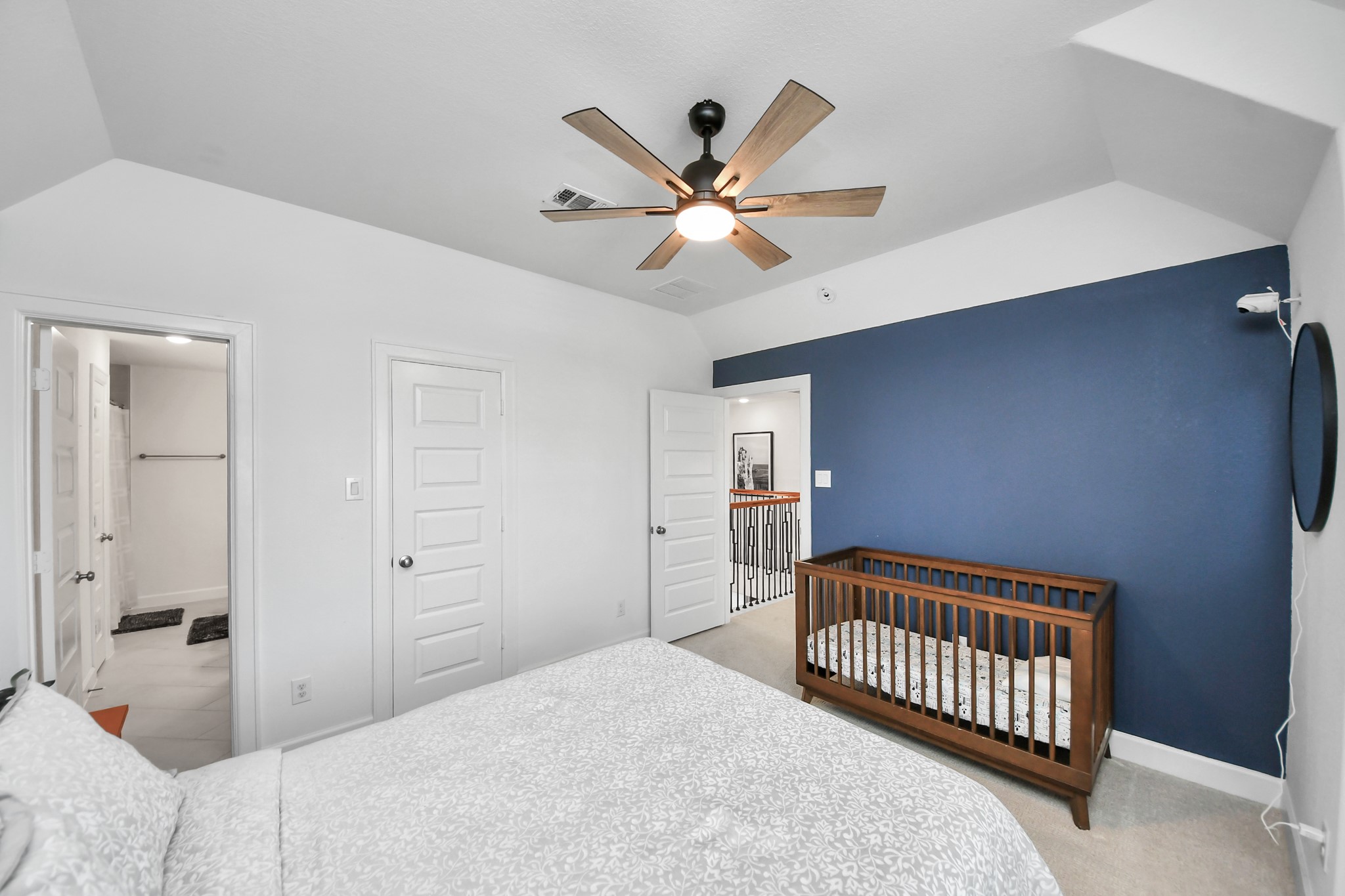 Charming bedroom featuring a cozy crib, elegant blue accent wall, and modern ceiling fan, with convenient access to an en-suite bathroom.