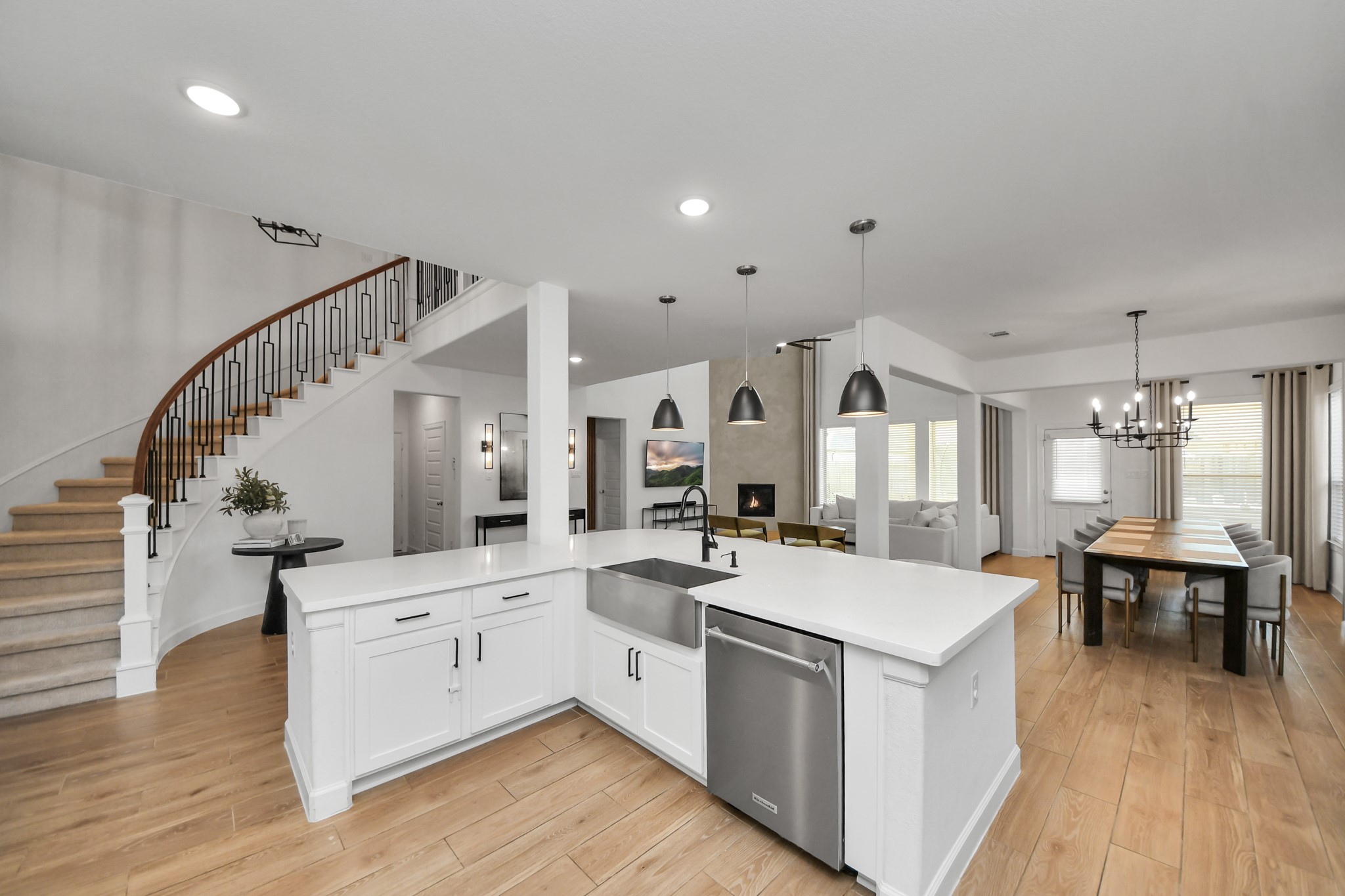 Elegant open-concept kitchen and dining area featuring sleek white cabinetry, modern pendant lighting, and a sweeping staircase, seamlessly connecting to a cozy living space with a fireplace.