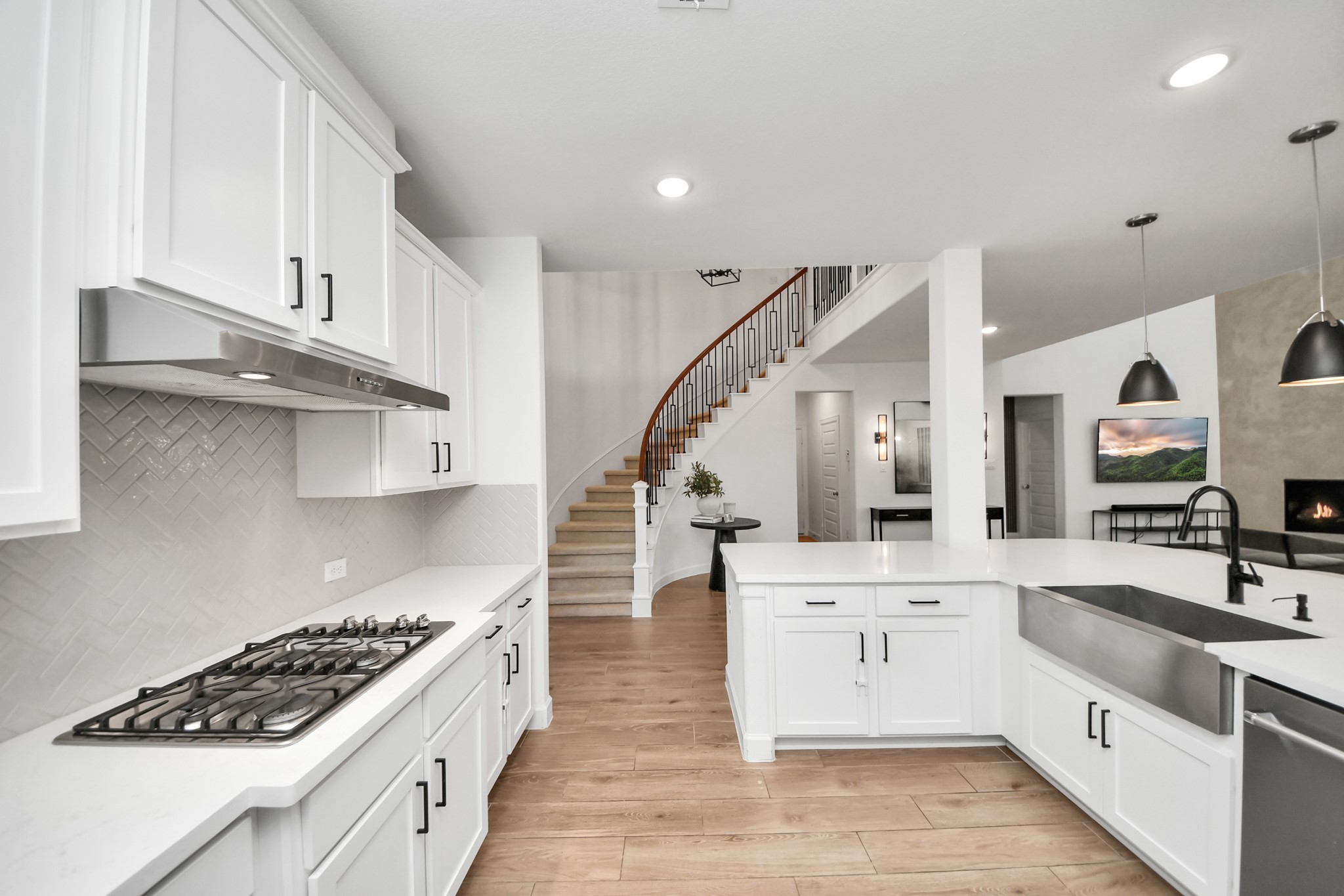 Spacious kitchen with sleek white cabinetry, a modern gas cooktop, and an elegant herringbone backsplash. The open layout flows seamlessly into the living area, highlighted by a curved staircase and warm wood flooring.