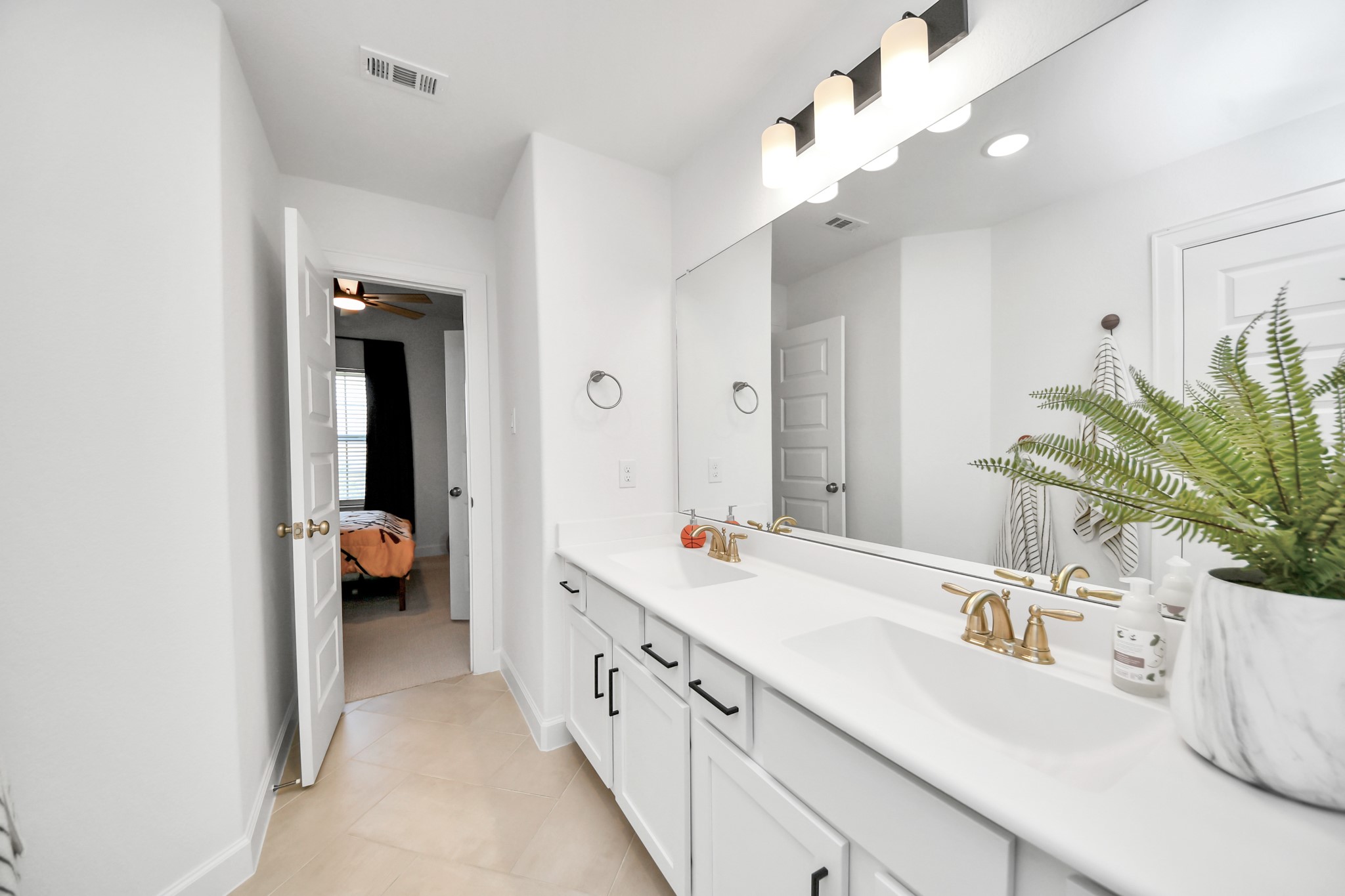 Bright and modern Jack and Jill bathroom featuring a double vanity with sleek gold fixtures, a large mirror, and ample storage, leading into a cozy bedroom.