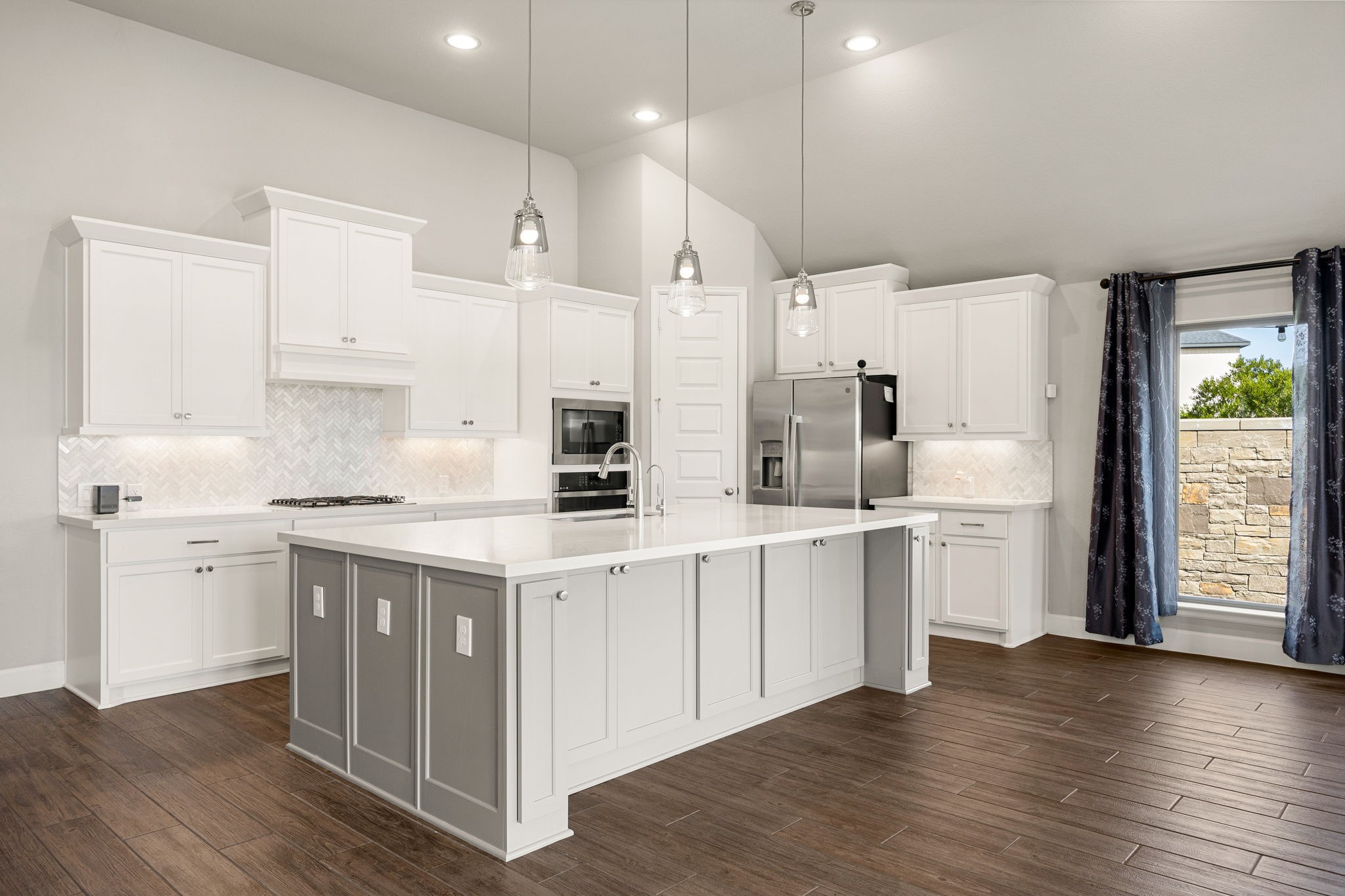 This view highlights the kitchen's beautiful proportions and impressive vaulted ceiling lines. The expansive quartz island features a two-tone accent finish, creating a sophisticated design anchor against the crisp white perimeter cabinets and built-in appliances.