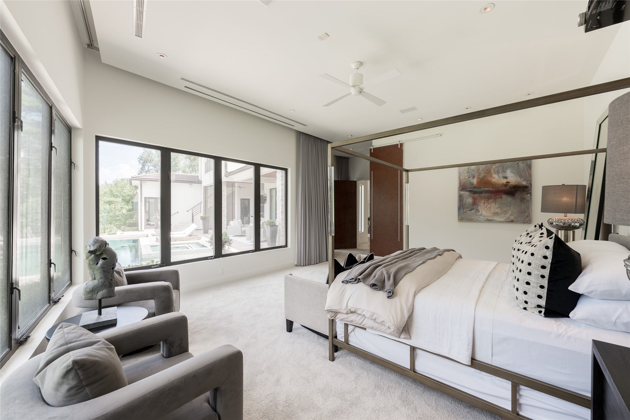 A second perspective of the primary suite showcases the corner windows and direct views of the pool and outdoor living areas. The rooms generous proportions provide space for a separate sitting area, while custom motorized shades offer privacy across the expansive windows.